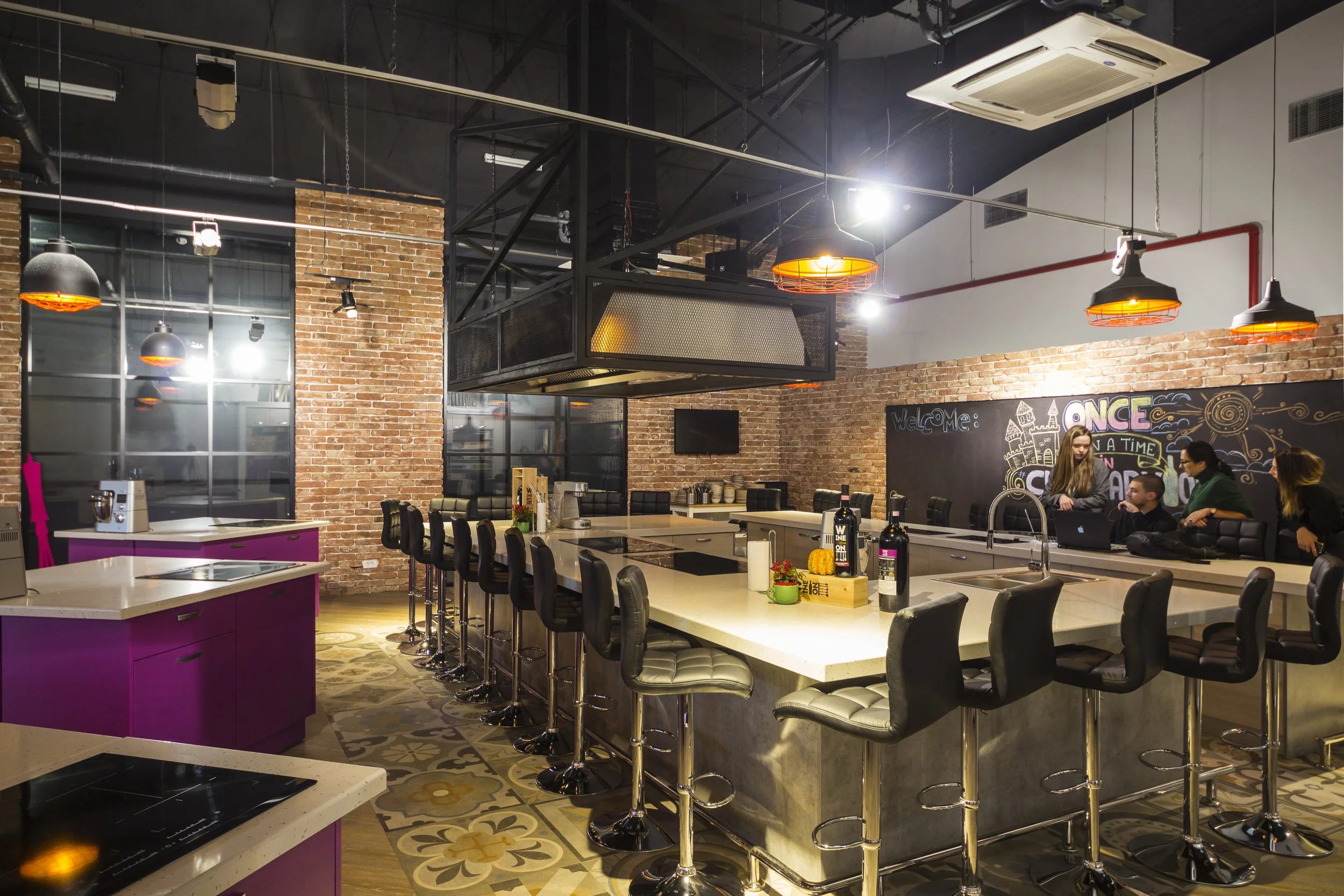Modern indoor cafe with a large central island counter surrounded by black bar stools, exposed brick walls, industrial lighting, and a black chalkboard wall with colorful writing and drawings.