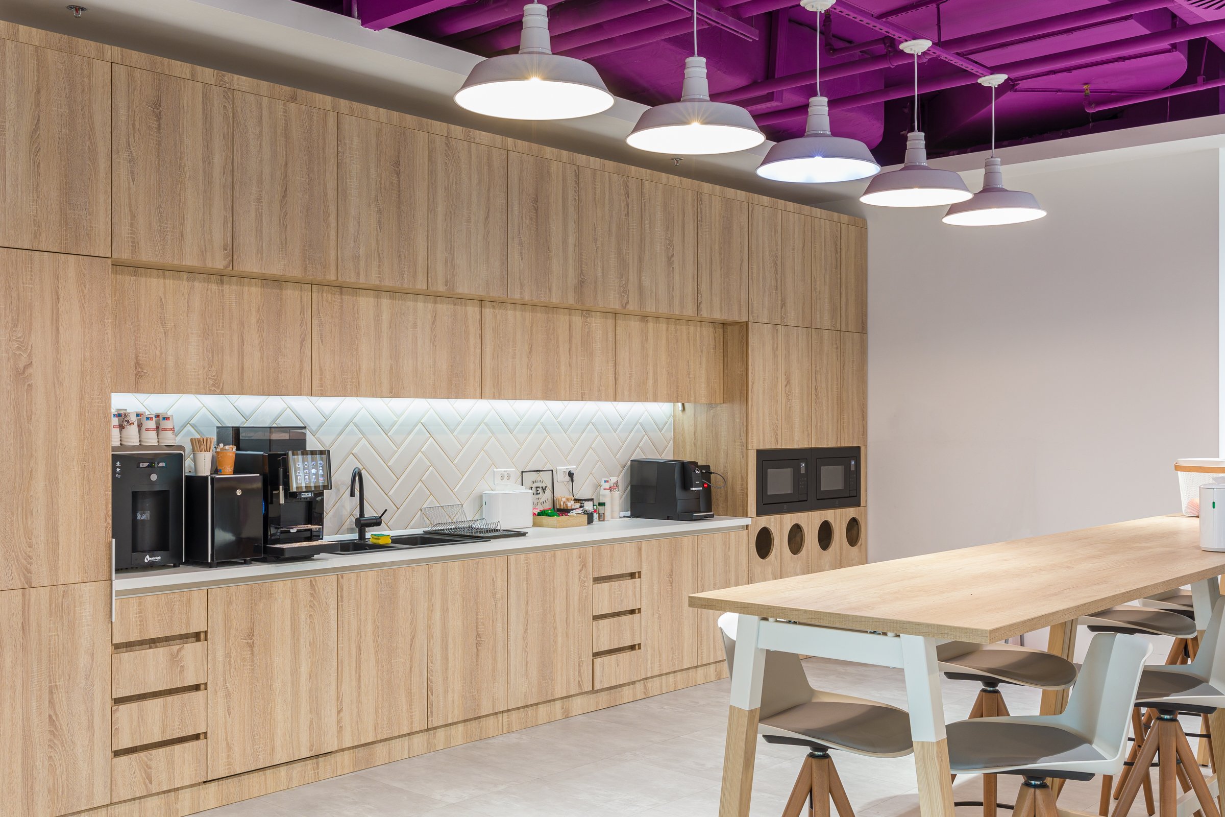 Modern kitchen with wooden cabinets, white countertop, coffee machine, microwave, and hanging pendant lights, adjacent to a wooden dining table with chairs.