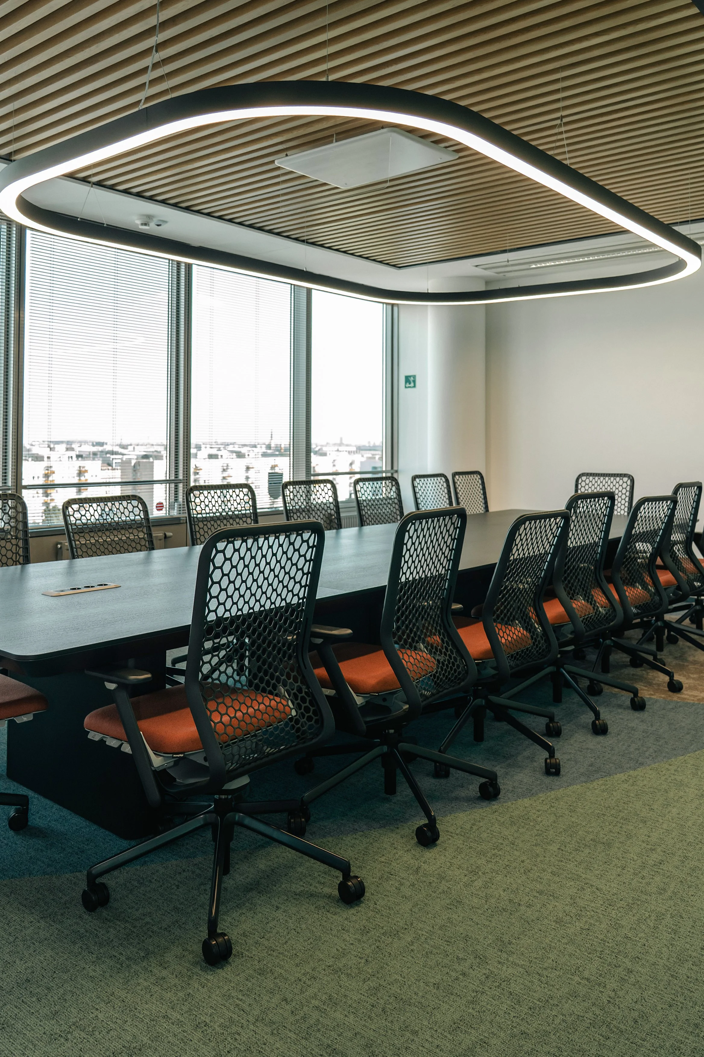Modern conference room with a long black table and multiple mesh-backed office chairs with orange seats, large windows with blinds, and a contemporary oval-shaped ceiling light fixture.