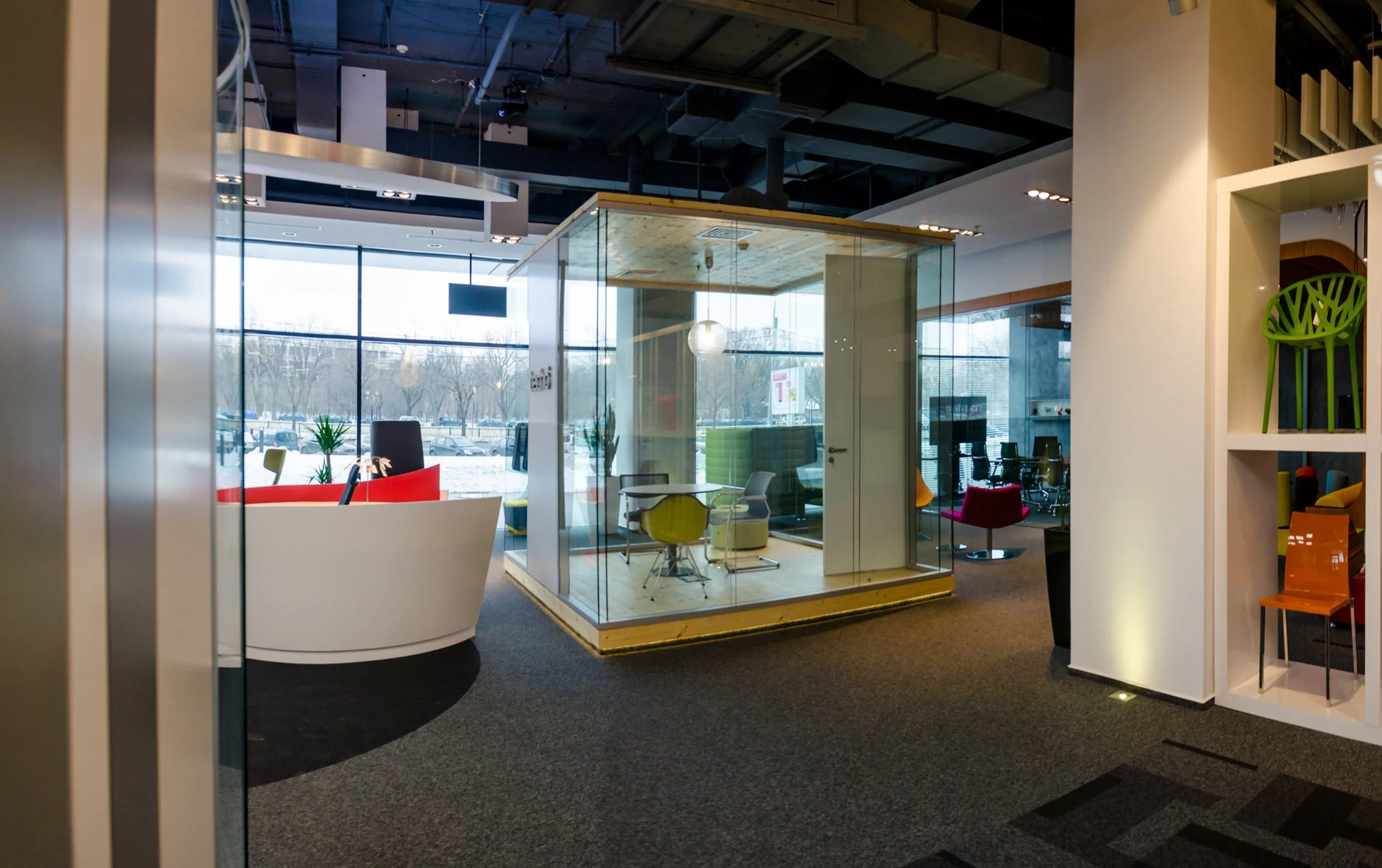 Modern office interior with glass-enclosed meeting room, colorful chairs, a white lounge area, and large windows showing a parking lot and trees outside.