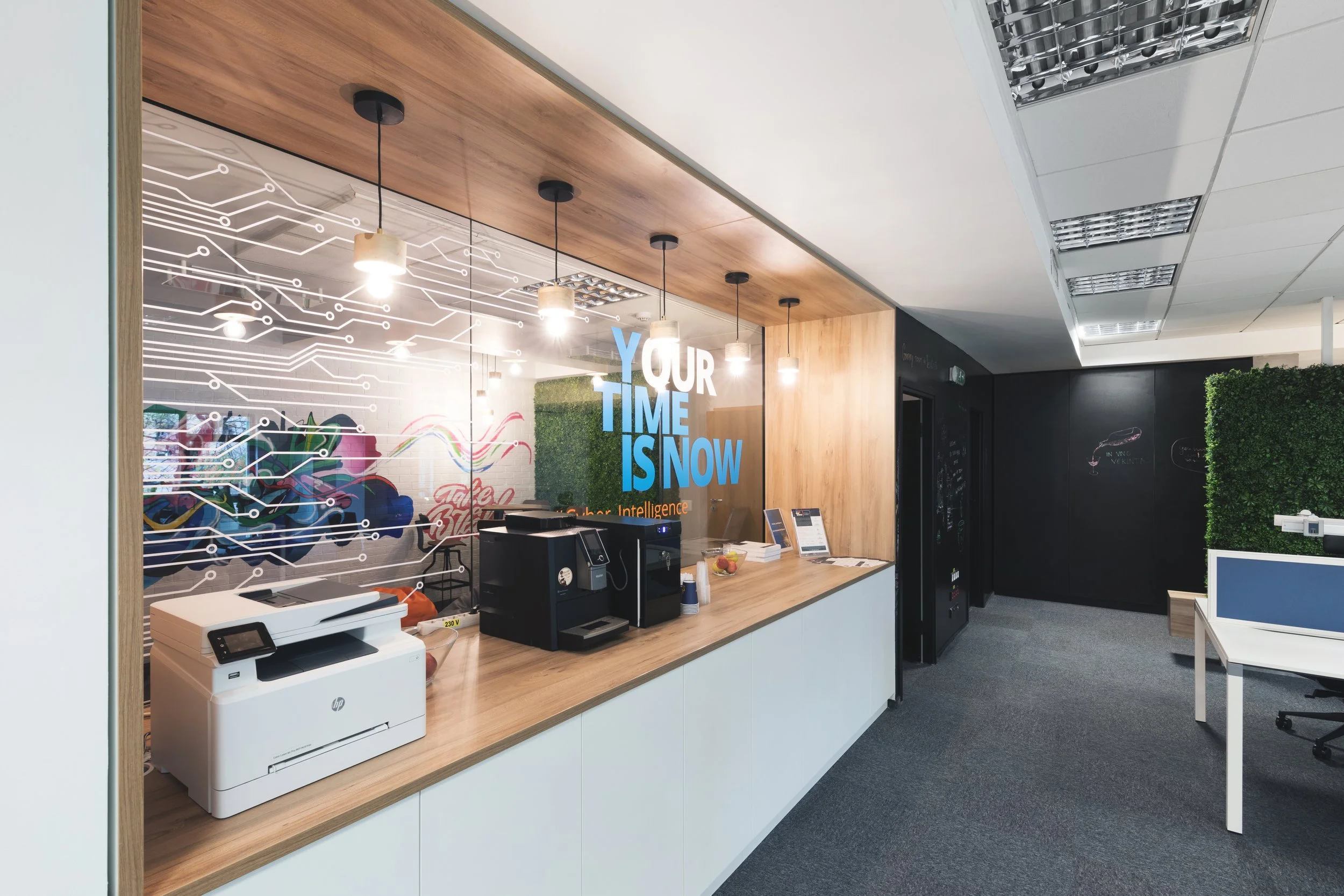 Office reception area with wooden counter, three hanging pendant lights, a printer, coffee machine, framed signs, and a glass wall with the text 'YOUR TIME IS NOW' in blue, and additional abstract and colorful decorations.