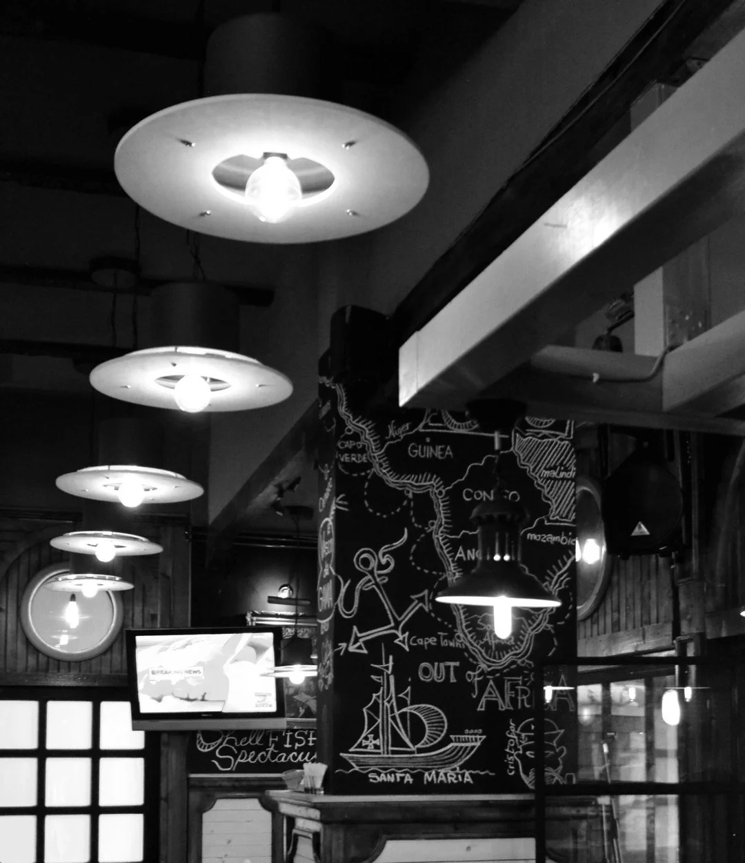 Black and white photo of a restaurant interior with overhead lights, a chalkboard with a map and writing, a TV screen, and wooden decor.