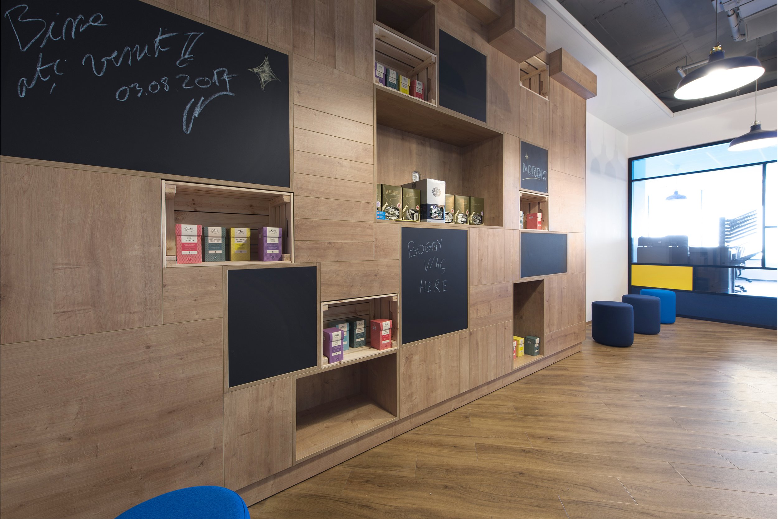 Modern interior with a large wooden shelving unit with black chalkboard sections and colorful boxed products, near a window with blue seating and stools.