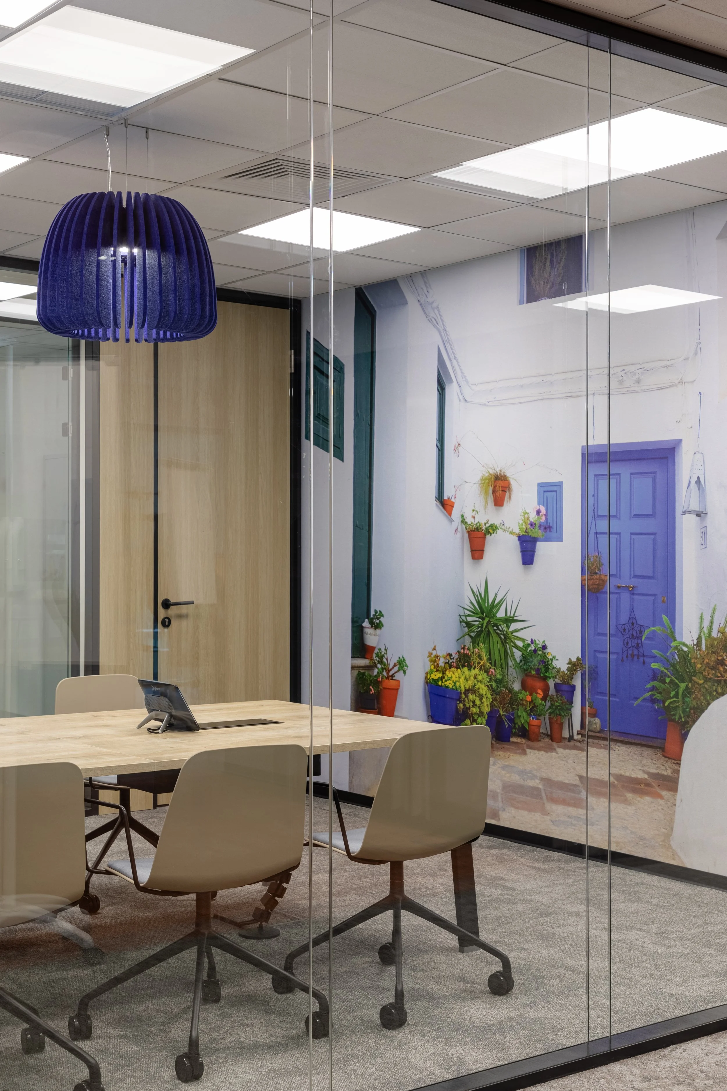 Office conference room with a wooden table, beige chairs, a blue pendant light, and a mural of a white building courtyard with potted plants and a purple door on the wall.