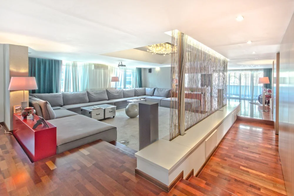 Spacious living room with gray sectional sofa, red accent table, pink lamps, large windows with curtains, and a glass partition with string beads, wooden floors, and natural light.