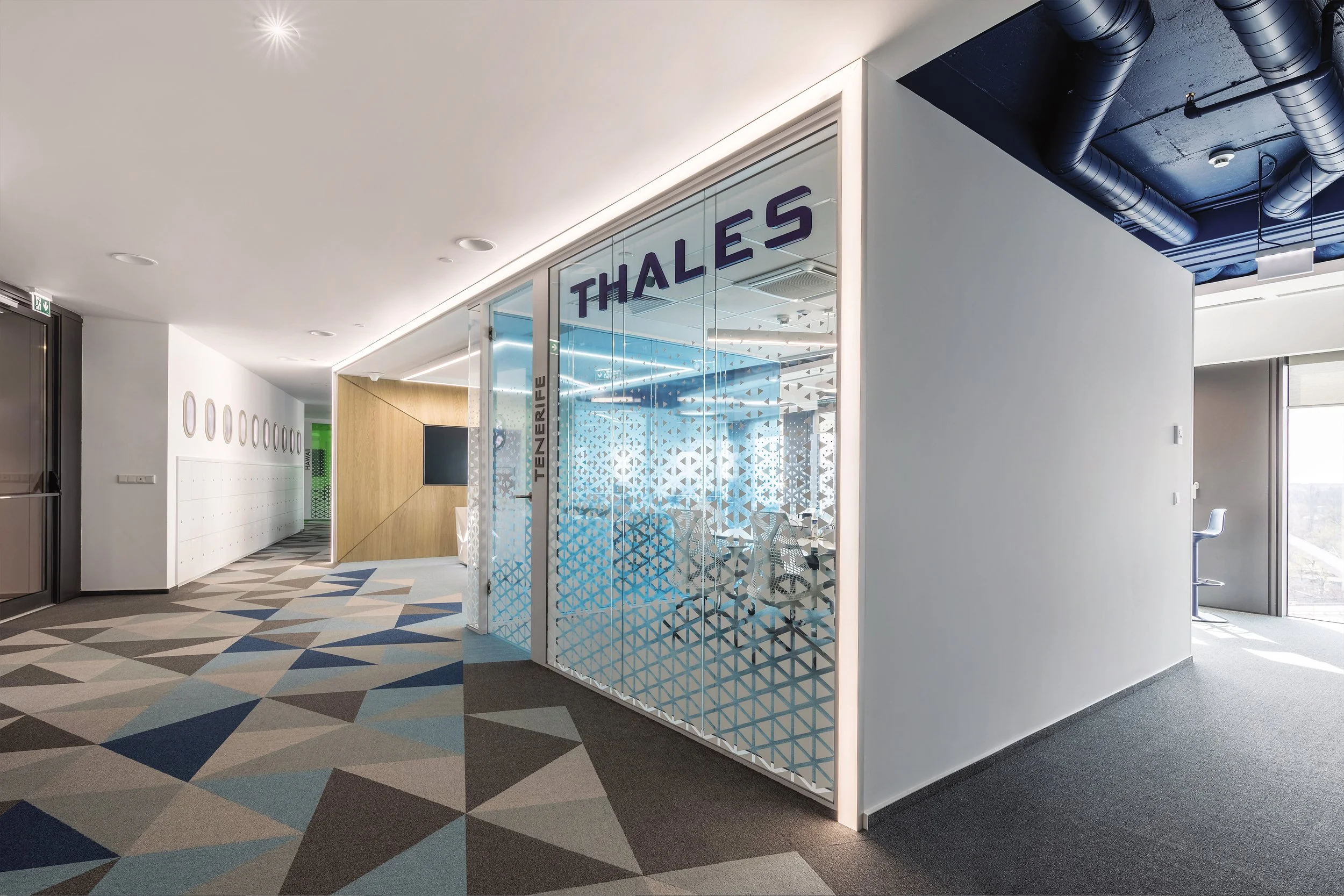 Modern office corridor with patterned carpet, glass meeting room with 'Thales' sign, and a seating area near large windows.