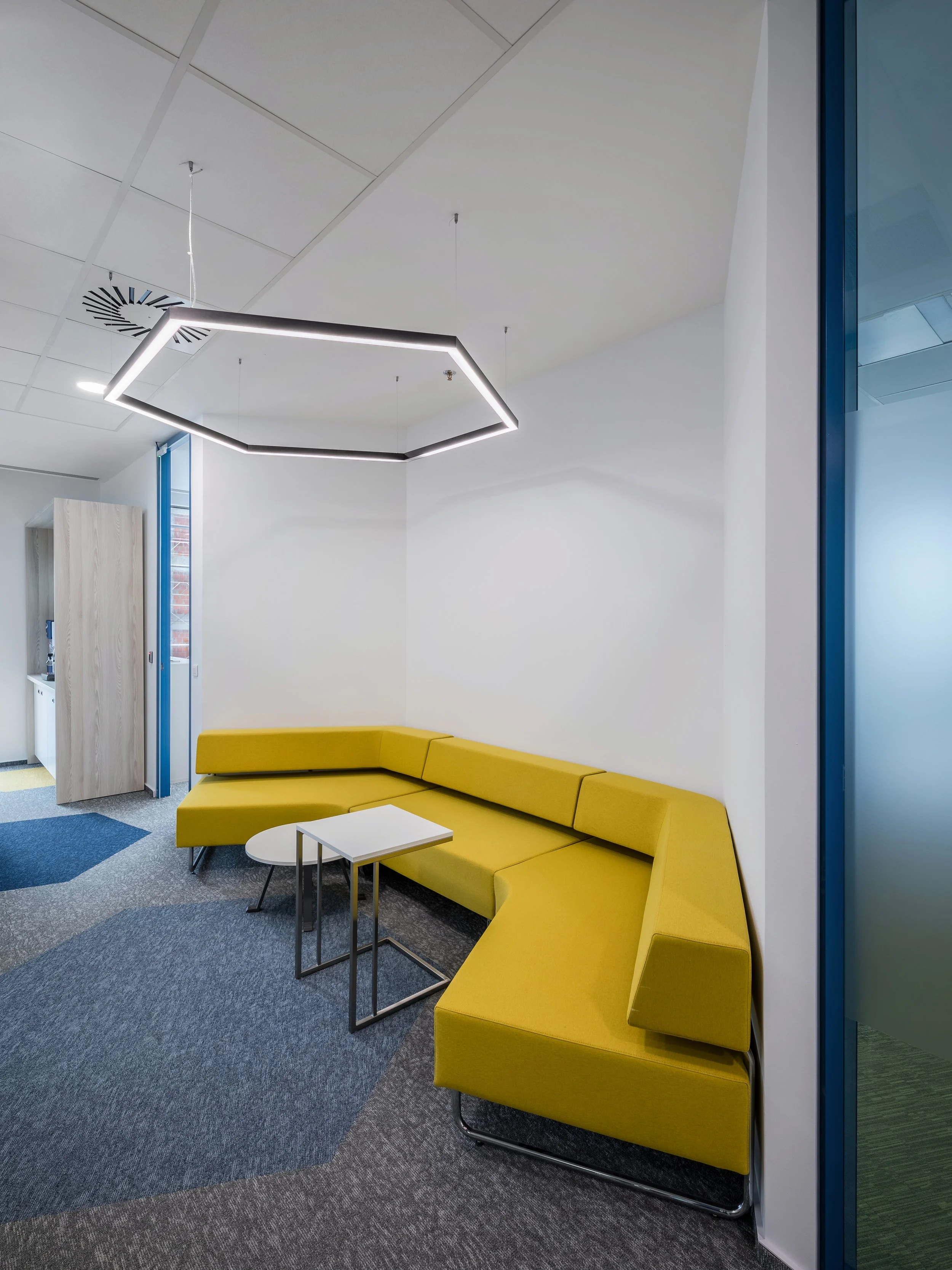 Modern office lounge with yellow sectional sofa, white tables, and geometric ceiling light fixture.