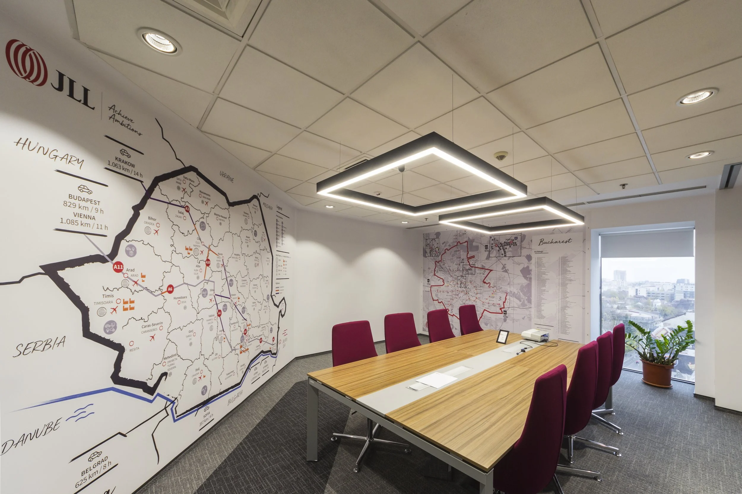 Conference room with a large wooden table surrounded by red chairs, a large wall map of Hungary and Romania, and a window with a city view, potted plant in the corner, and modern ceiling lighting.
