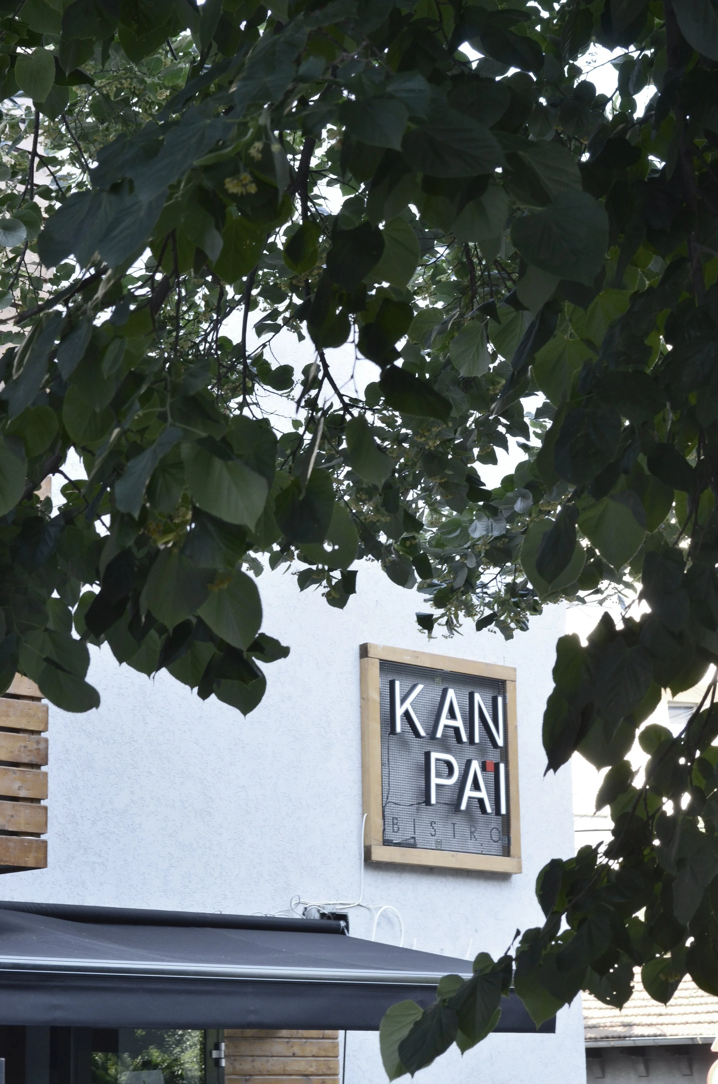 Sign for KAN PA'I BISTRO on the wall of a building, partially obscured by green leaves and branches.