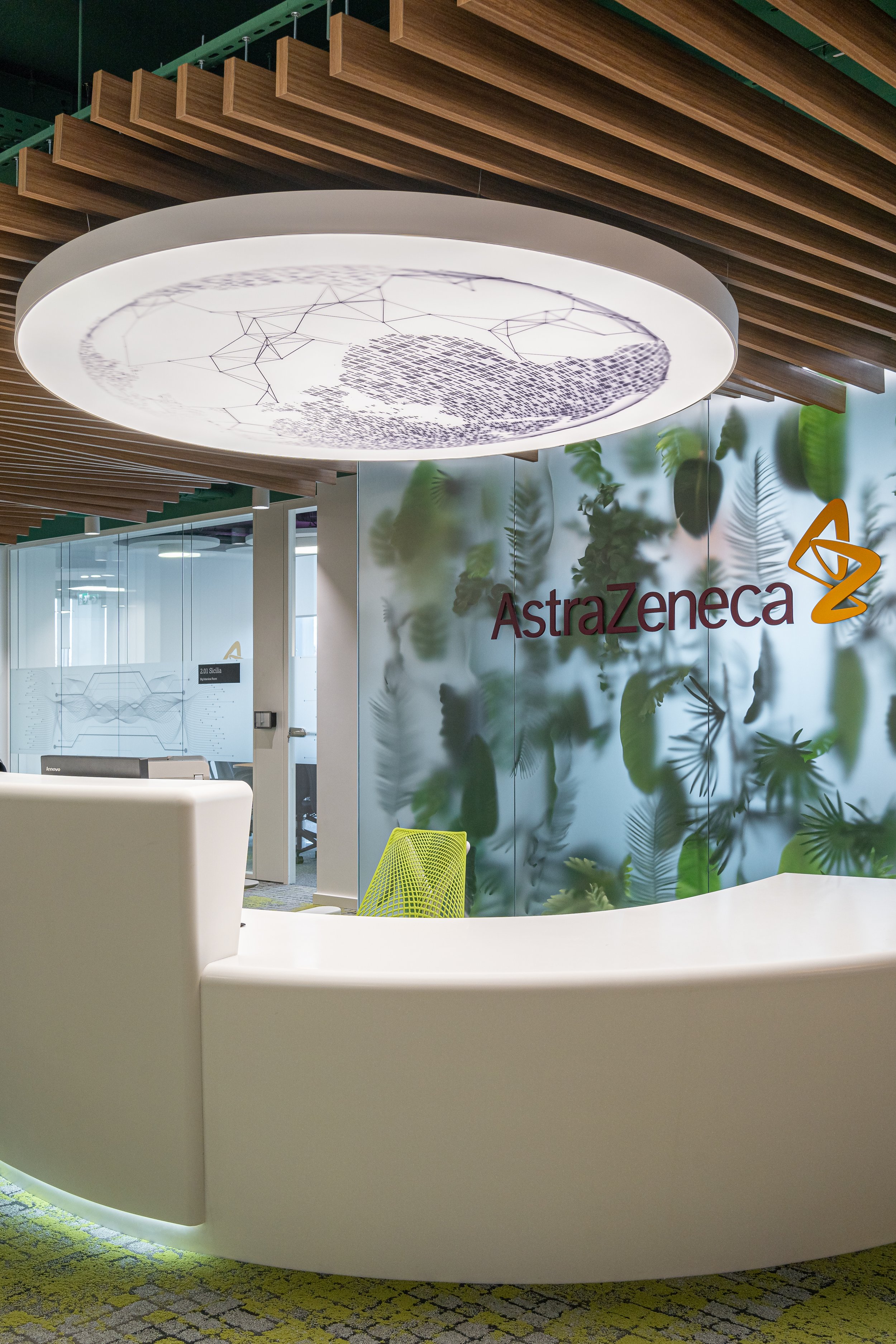 Modern reception area of AstraZeneca with a curved white desk, a green mesh office chair, a large illuminated ceiling fixture, and a wall with the AstraZeneca logo and a green leafy pattern.