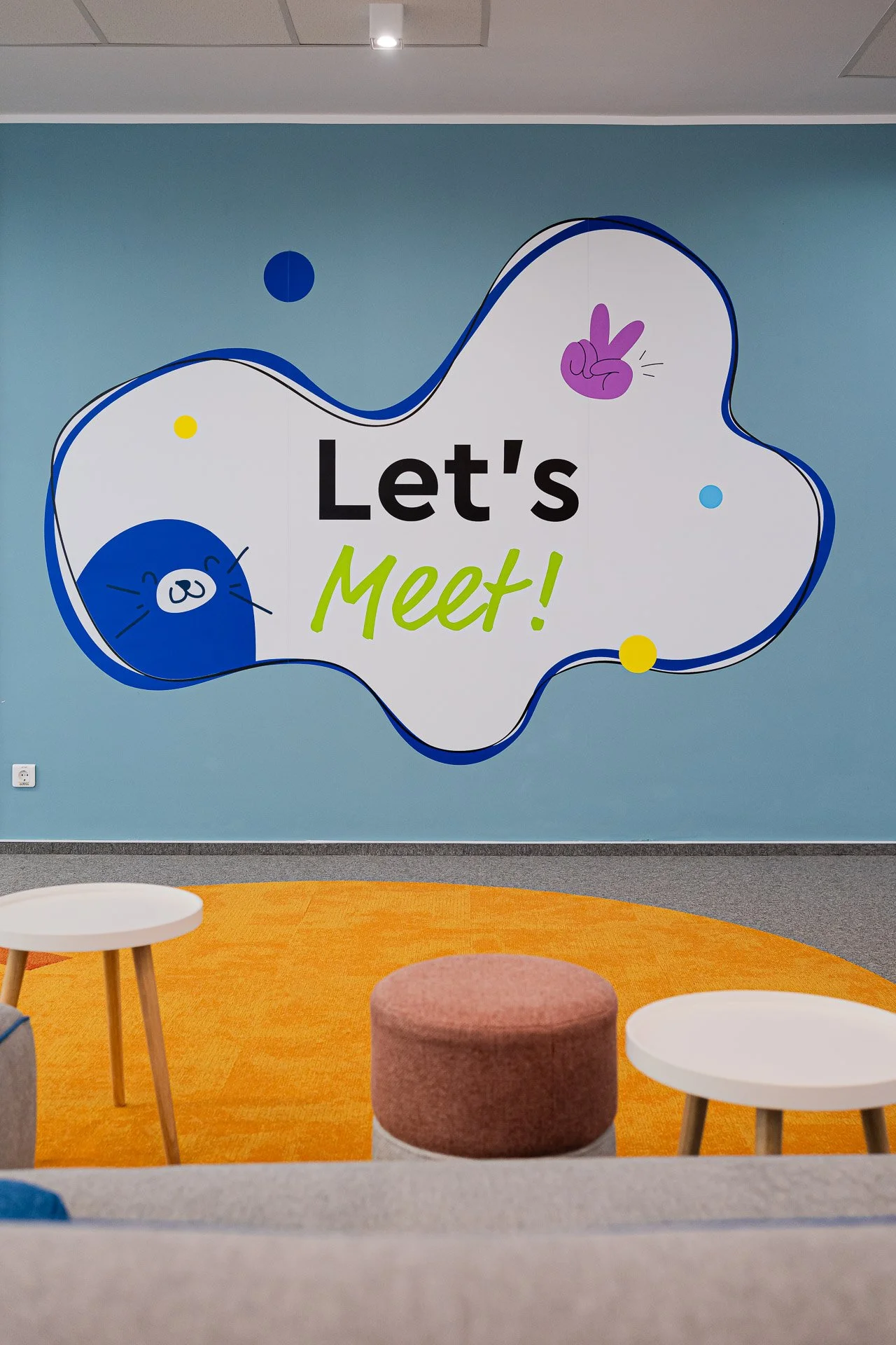 Colorful office meeting room with a wall sign saying "Let's Meet!" decorated with cartoon illustrations of a bear and a hand making a peace sign, with small colorful dots around. The room has a round orange rug, a pink cushioned stool, and two white circular tables.