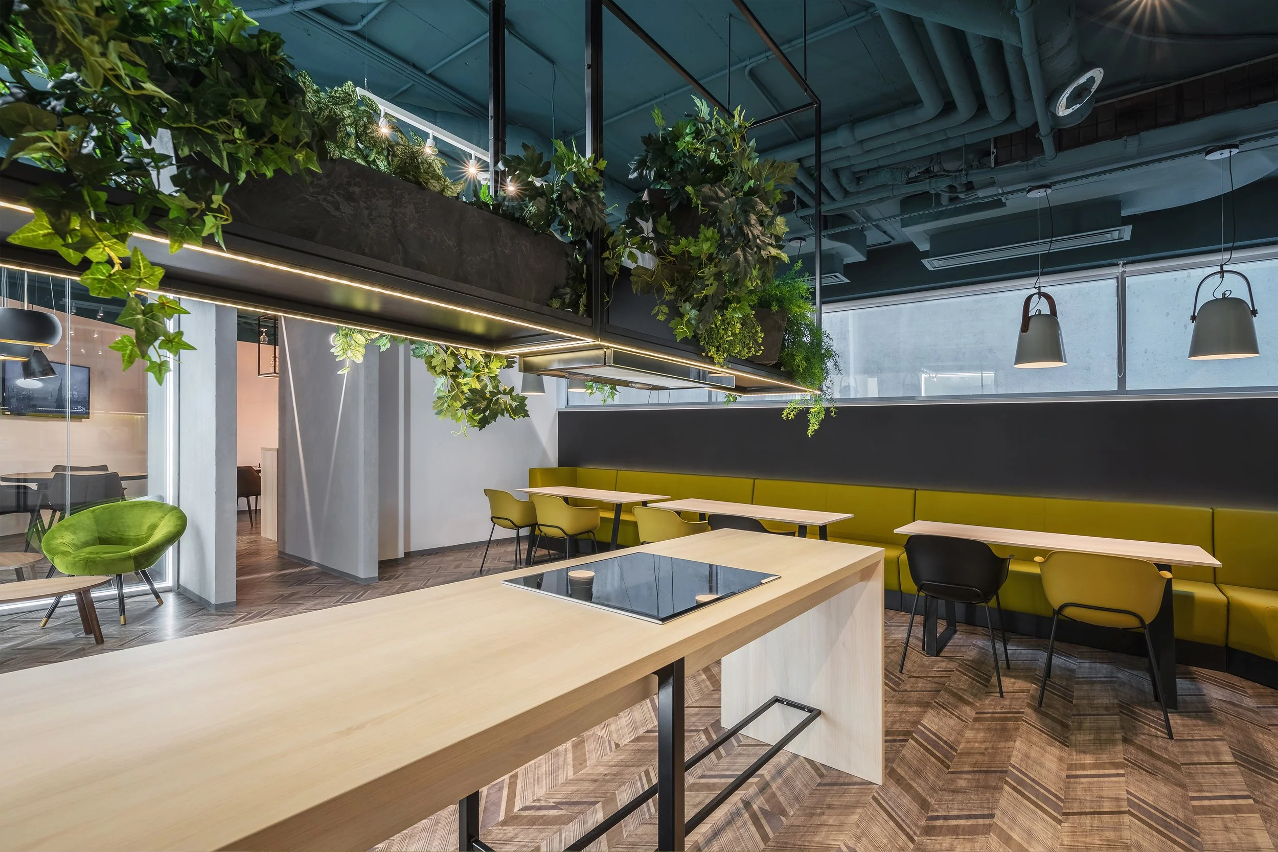 Modern indoor space with a wooden table, green seating, hanging plants, pendant lights, and large windows in the background.