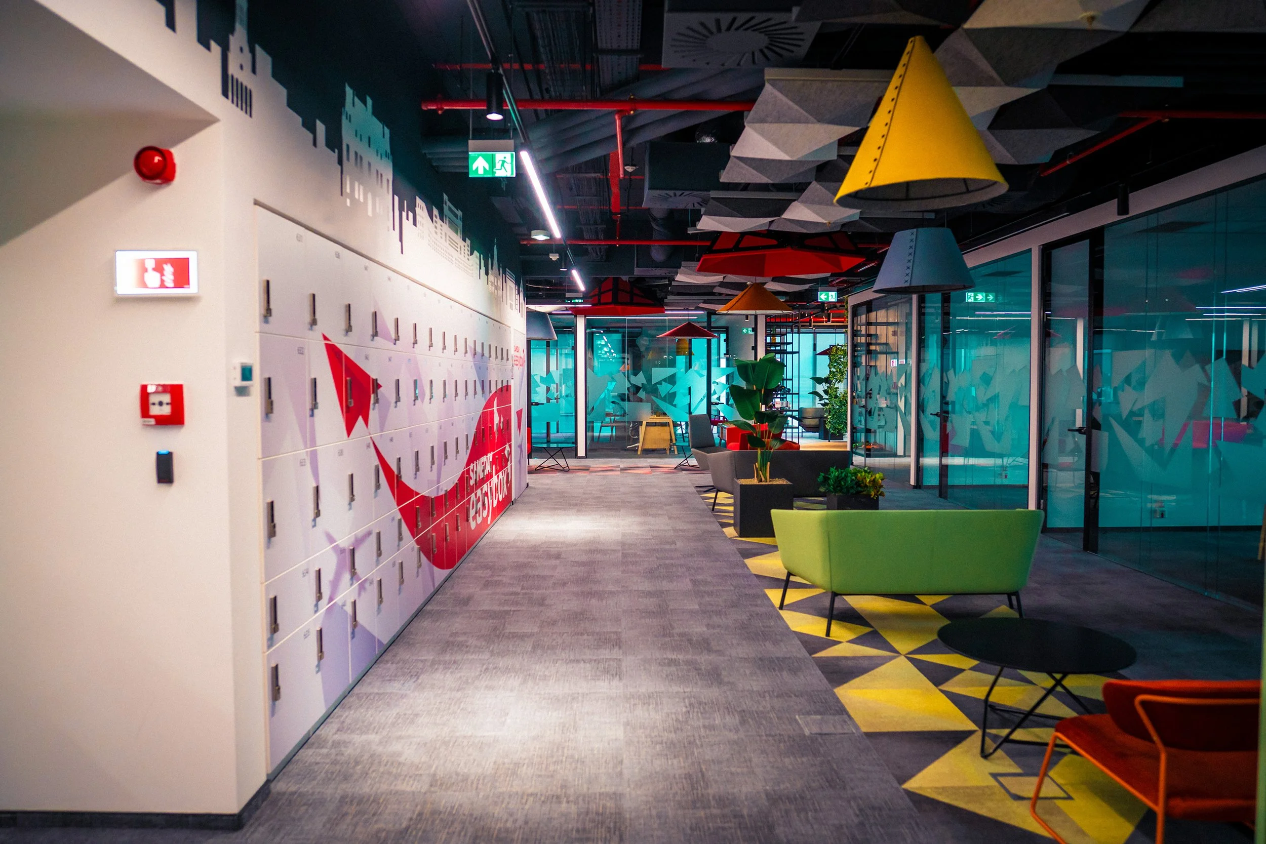 Modern office lounge with colorful furniture, plants, lockers, and geometric ceiling decor.