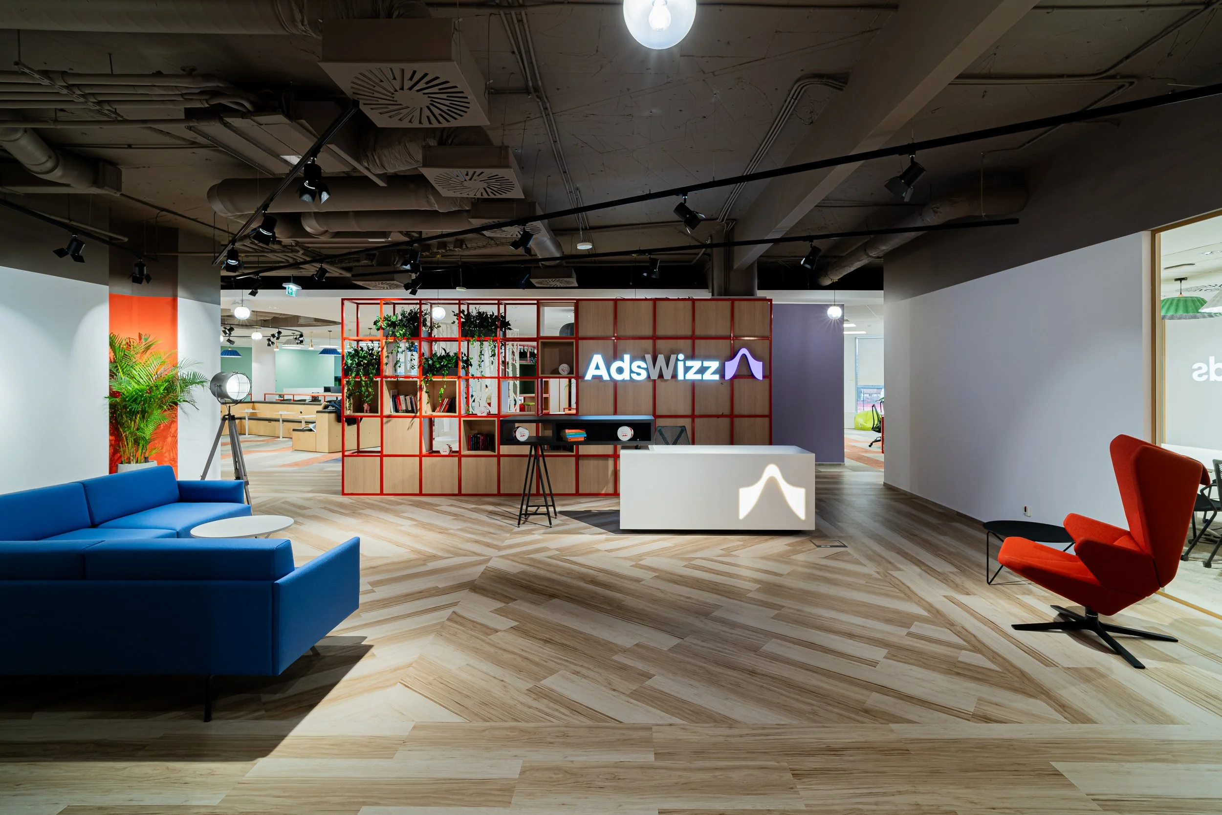 Modern office reception area with a wooden floor, blue sofa, red chair, green plants, and a sign that reads 'AdsWizz' on a red grid partition wall.