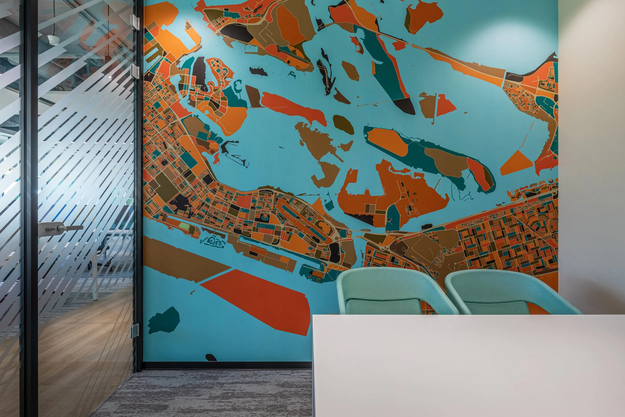 A large, colorful map mural on the wall of an office conference room, depicting a city and waterways in a stylized, abstract manner. There are three green chairs in front of the map and a white table.