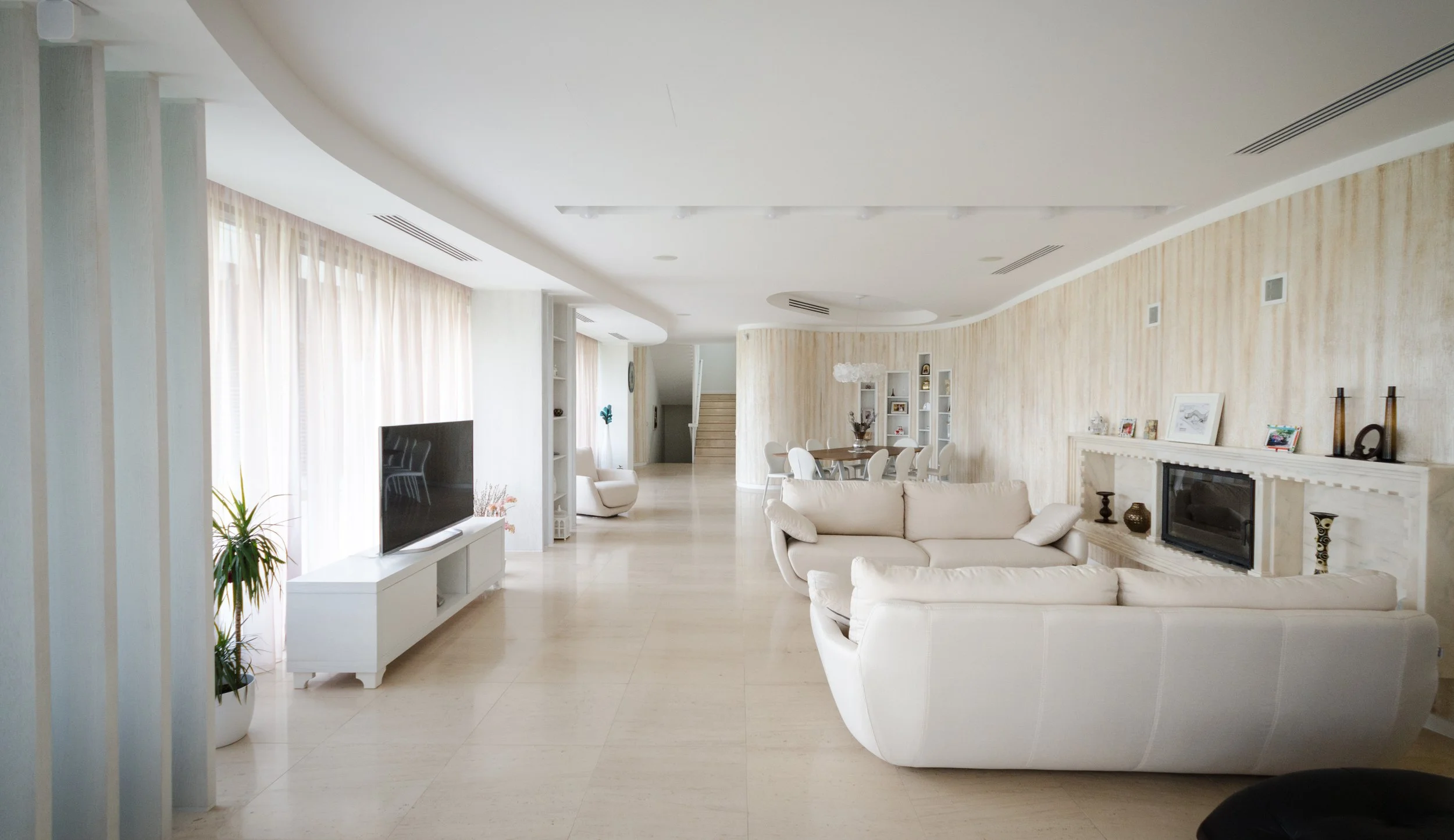 Spacious living room with cream-colored sofas, a flat-screen TV on a white stand, and a fireplace with decorative objects, pastel-colored walls, and large windows with light curtains.