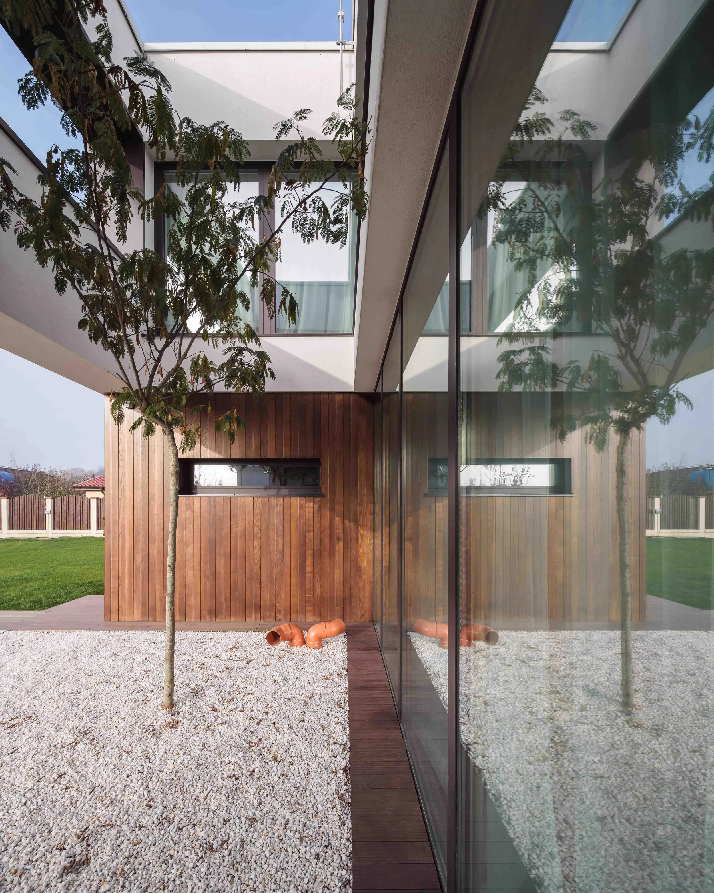 Modern house with a courtyard featuring a tree, gravel ground, and a wooden wall, with large glass window reflecting the tree.