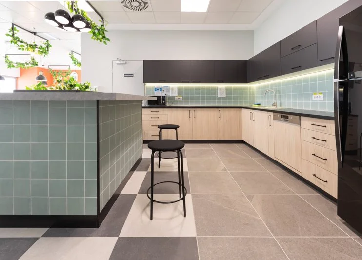Modern kitchen with black upper cabinets, light wood lower cabinets, green tiled backsplash, black countertop, and a breakfast bar with two black stools. There are plants, a microwave, and a refrigerator visible.