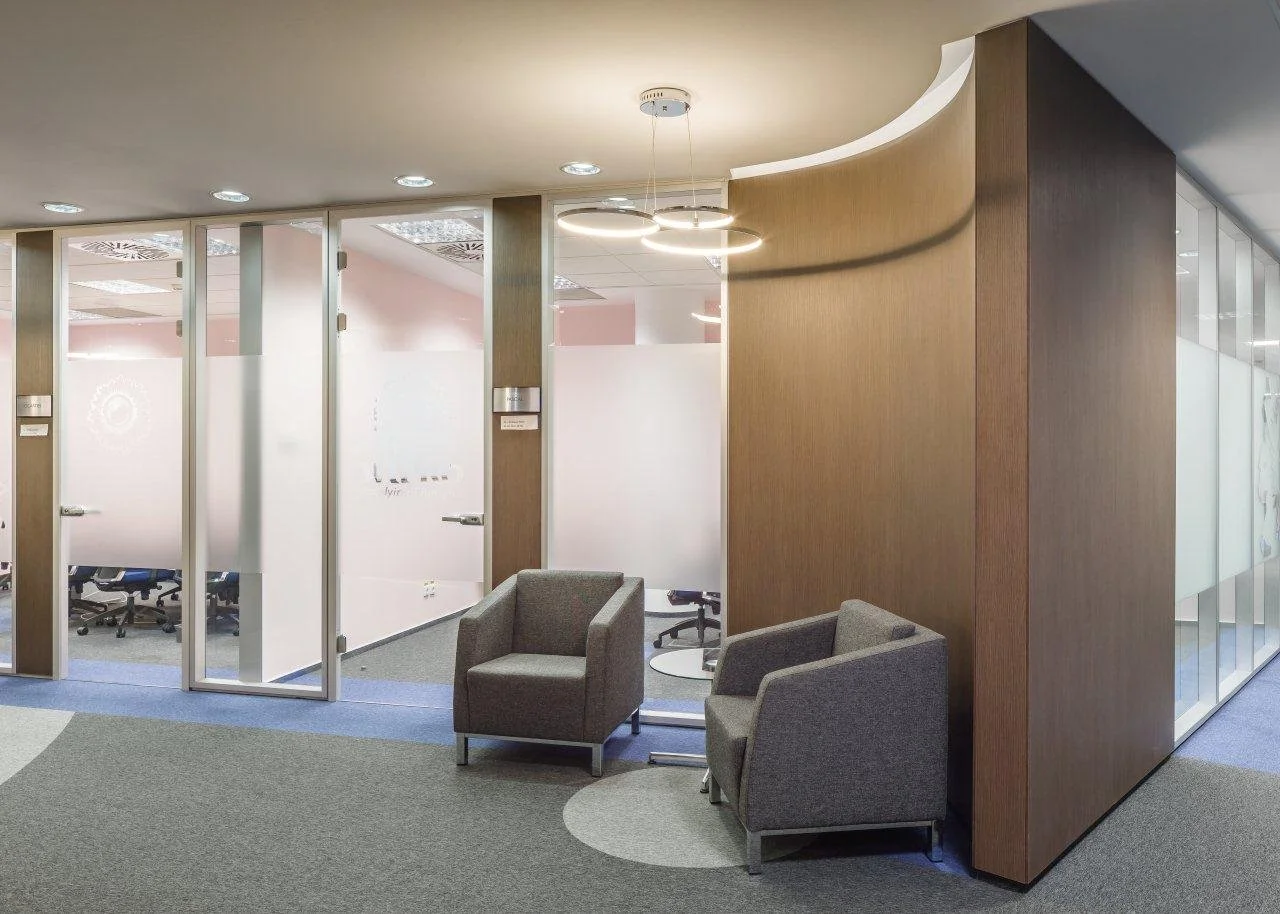 Modern office lounge area with two gray armchairs, a small glass table, glass walls with frosted signs, and a distinctive ceiling light fixture.