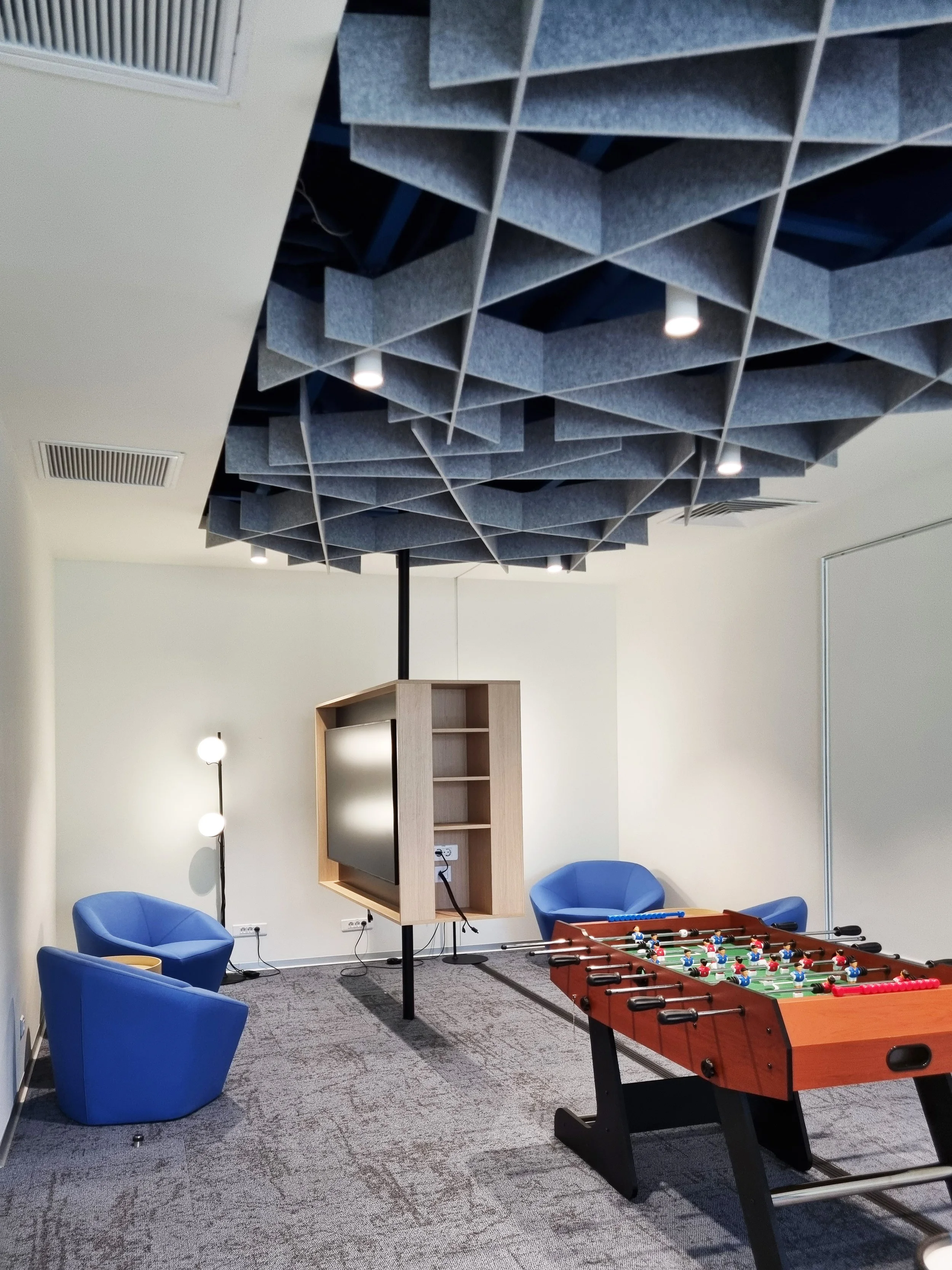 A modern lounge with two blue armchairs, a foosball table, a flat screen TV on a wooden stand, a standing floor lamp with two round bulbs, and acoustic panels on the ceiling.