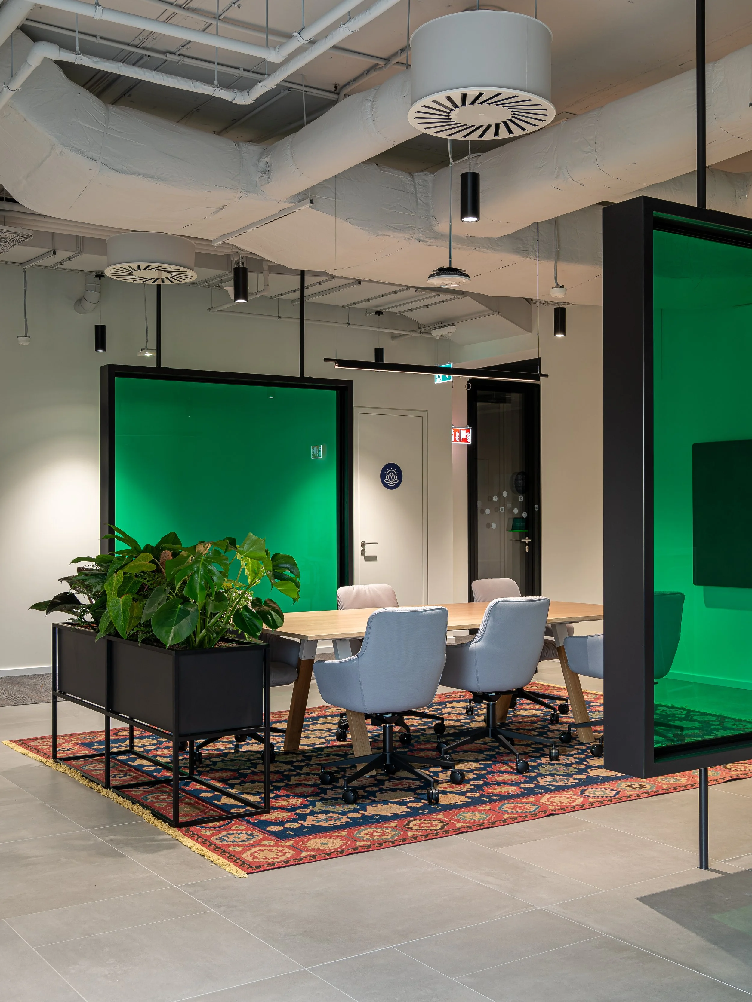 Modern office conference room with a wooden table, four white swivel chairs, a decorative carpet, large green panels, and a black plant stand with a leafy green plant, with exposed ceiling pipes and lighting.