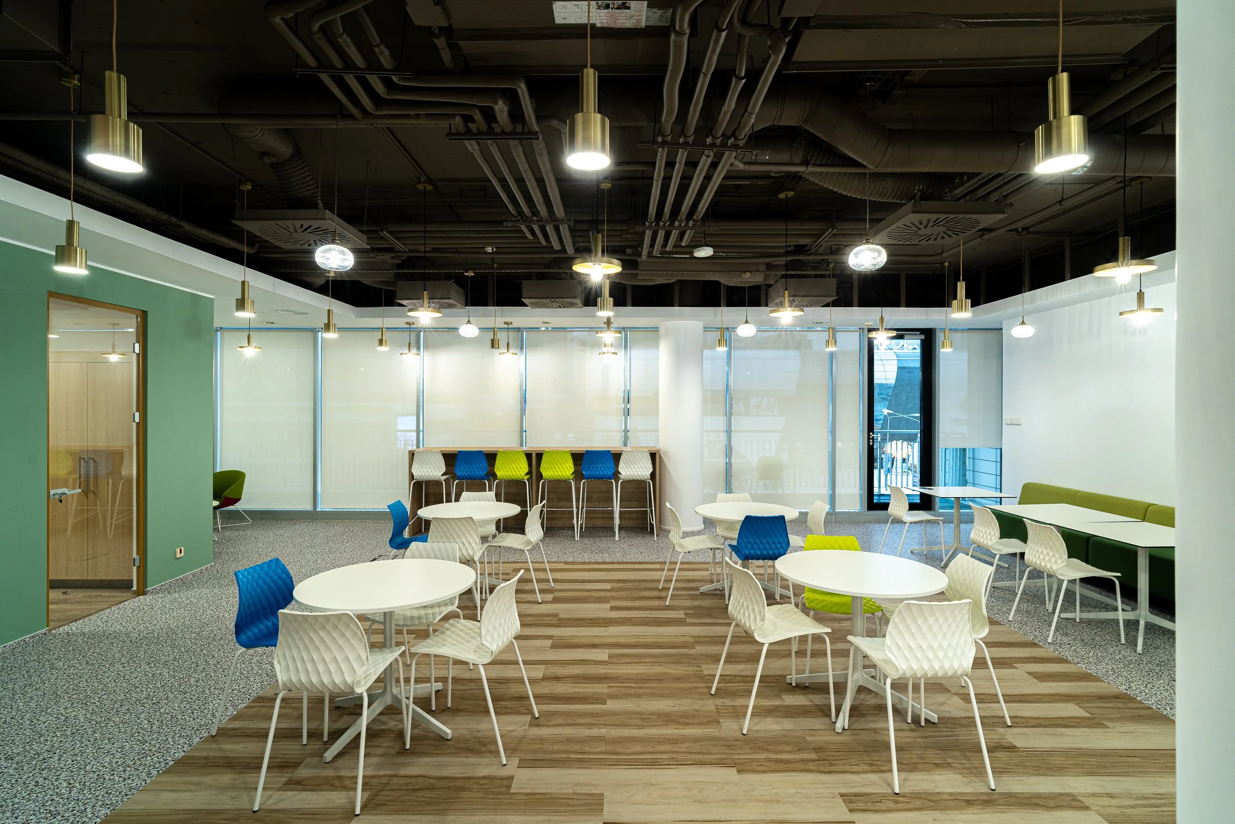 Modern communal workspace with round white tables, white and blue chairs, and a green bench along the wall, illuminated by hanging white and metallic lights.