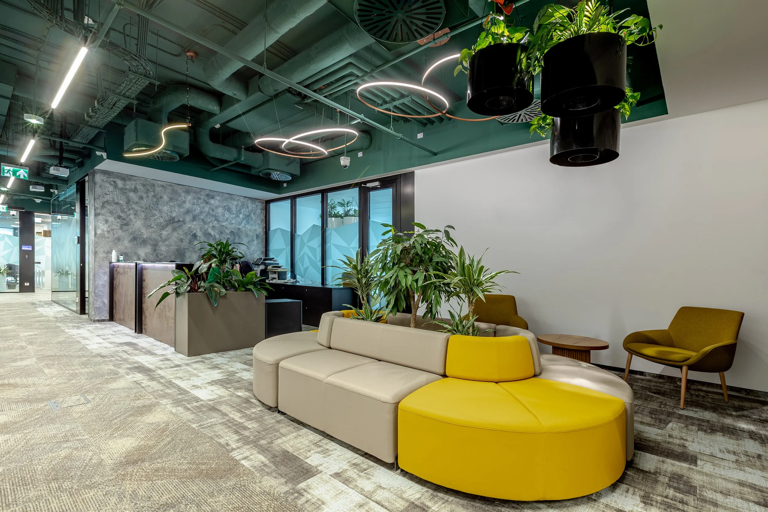 Modern office lounge area with beige and yellow sectional sofa, green potted plants, and mustard-colored chairs, illuminated by decorative ring and linear pendant lights, with gray textured wall and glass window in the background.