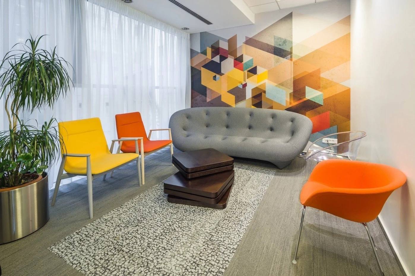 Modern office waiting area with colorful chairs, a gray tufted sofa, a wooden coffee table, a potted plant, a geometric patterned wall art, sheer white curtains, and a light-colored carpet.