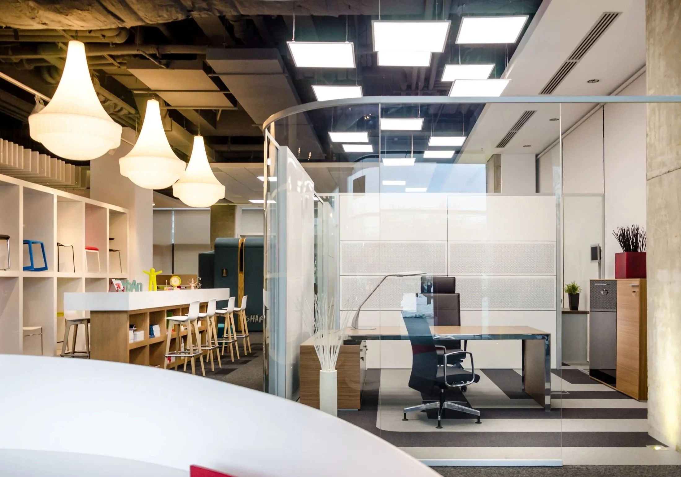 Modern office space with glass-walled private office, white and wooden furniture, black office chair, decorative plants, and hanging pendant lamps.
