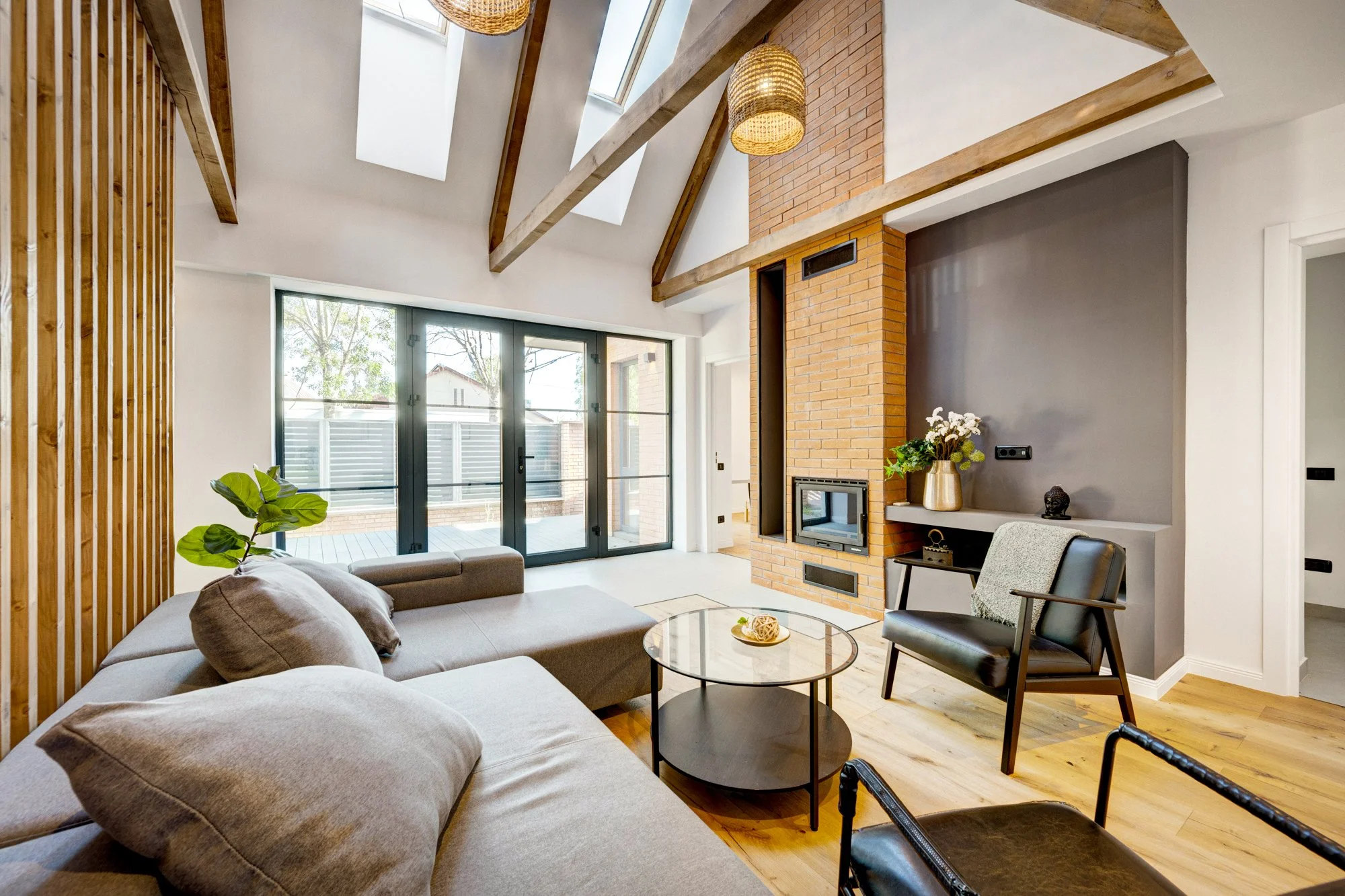 Modern living room with sofa, armchair, coffee table, fireplace, and large glass doors leading to outdoor patio, featuring wood beams, skylights, and decorative indoor plants.