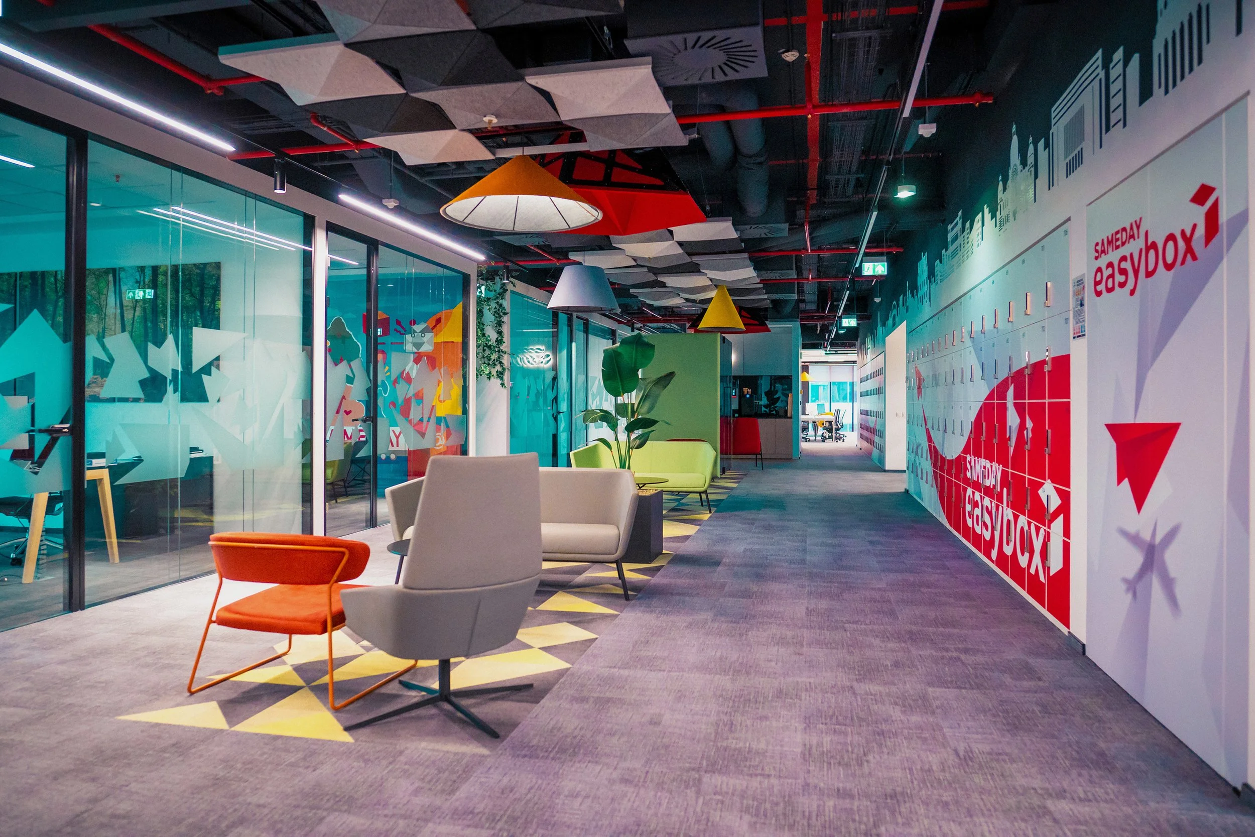 Modern office lounge area with colorful chairs, sofas, and hanging lamps. Glass walls with abstract designs, potted plants, and a mural on the wall reading "Sameday Easybox."