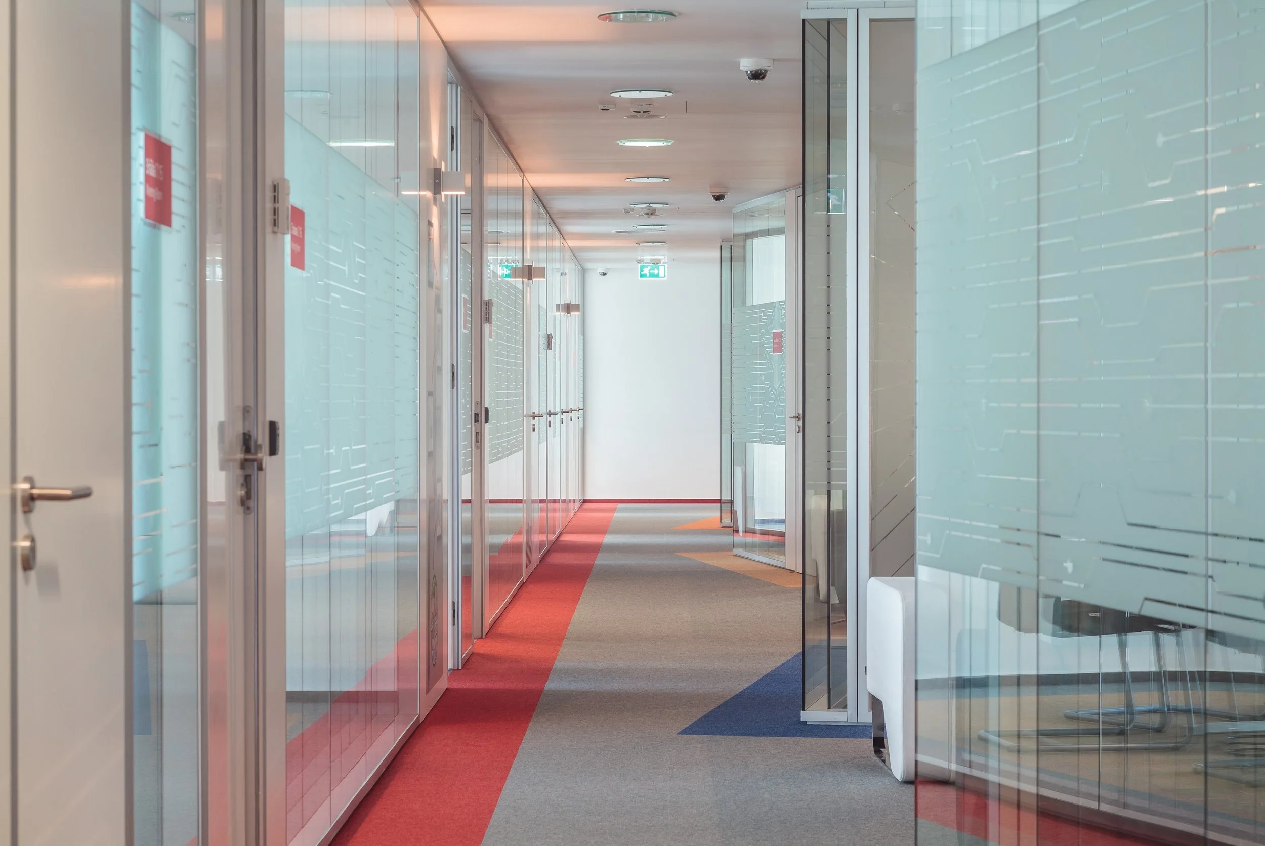 Empty office hallway with glass walls, closed glass doors, orange and blue carpet, white ceiling, and a visible exit sign at the end.