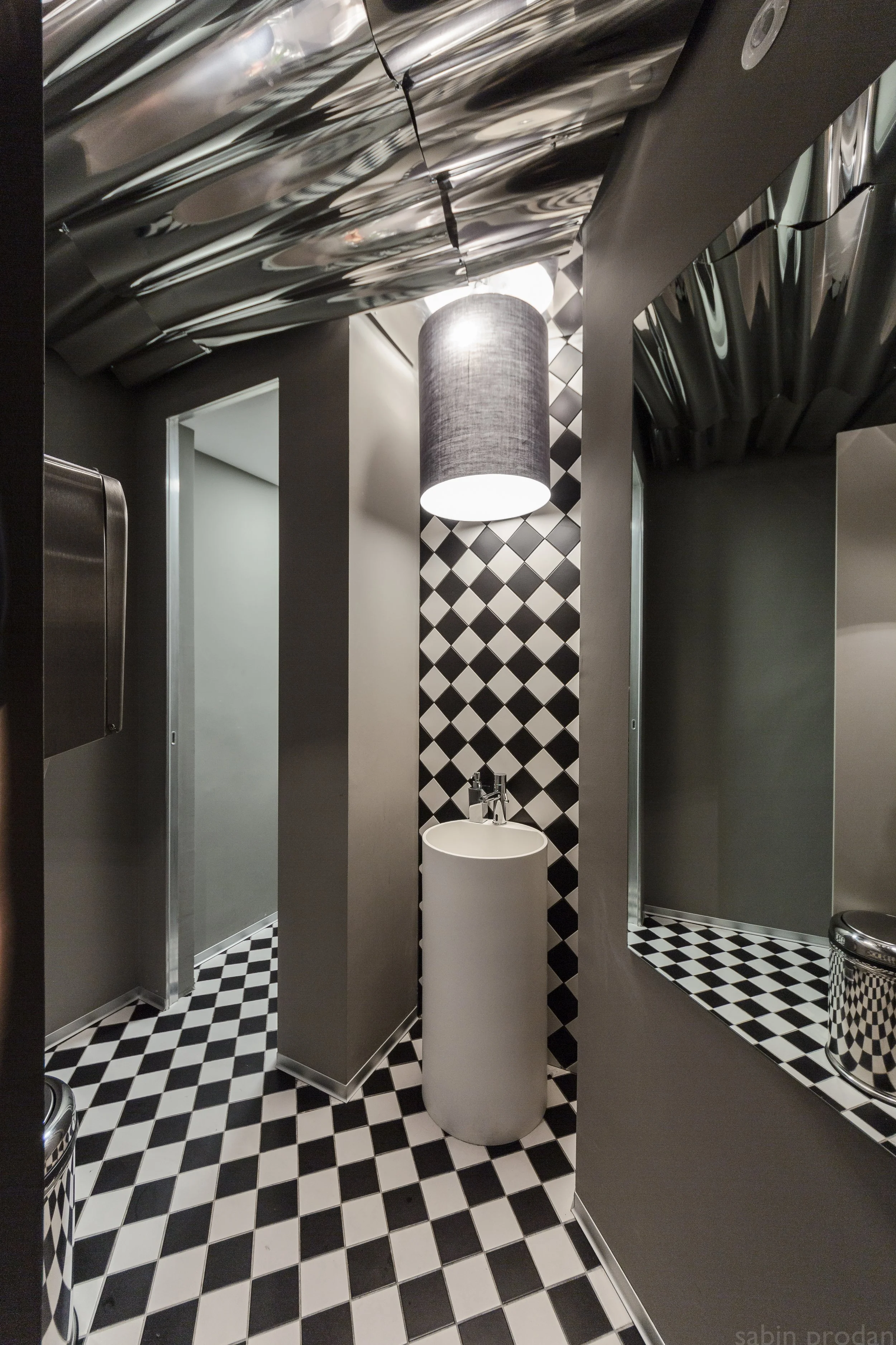A modern bathroom with black and white checkered tiles on the floor and a wall behind a small white cylindrical sink. There is a large mirror on the right, reflective metallic ceiling panels, a curtain-covered light fixture hanging from the ceiling, and part of a stainless steel trash can visible in the lower right corner.
