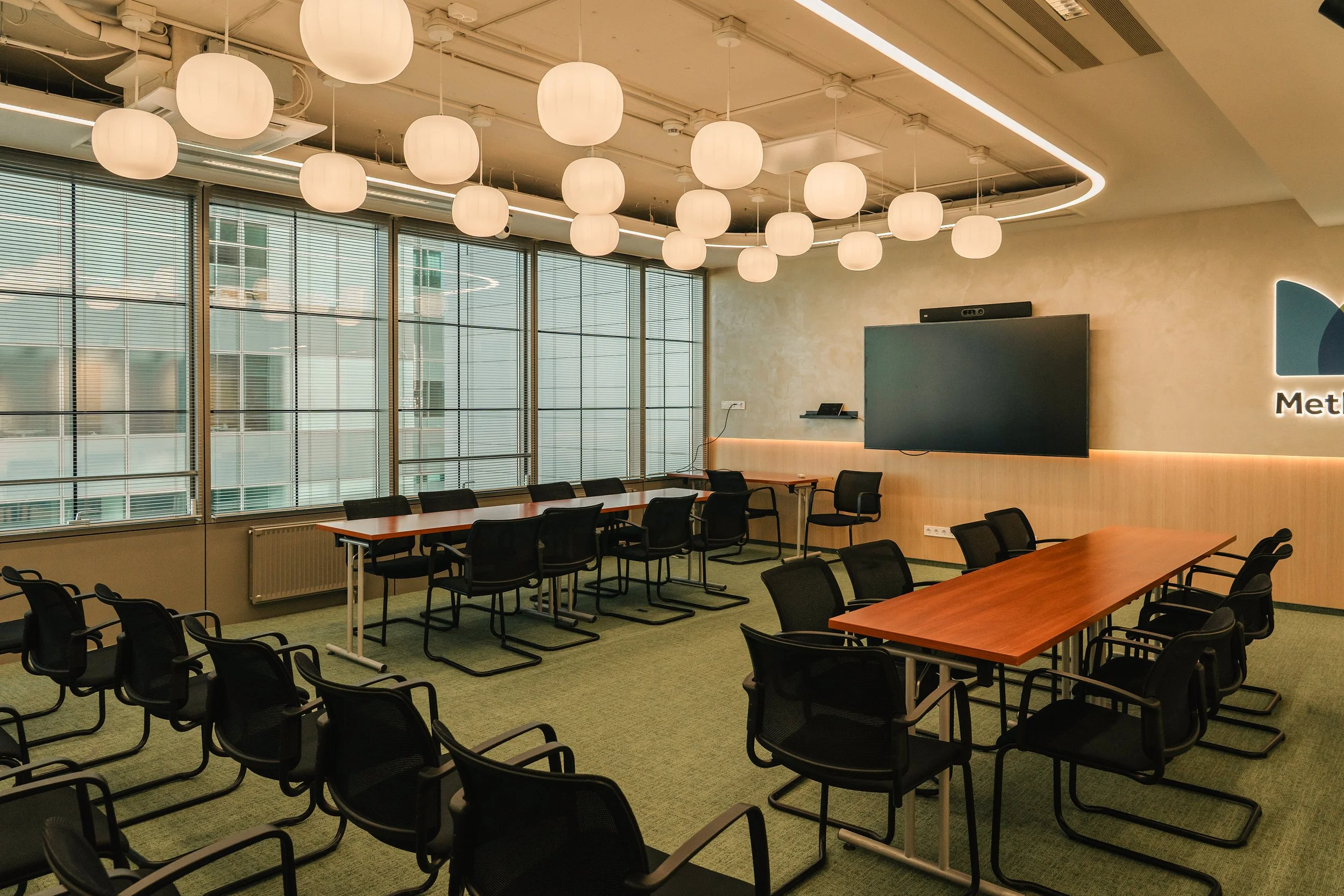 Modern conference room with black chairs, long tables, large windows with blinds, hanging white lantern lights, a flat screen TV on the wall, and a logo that says 'Met'.