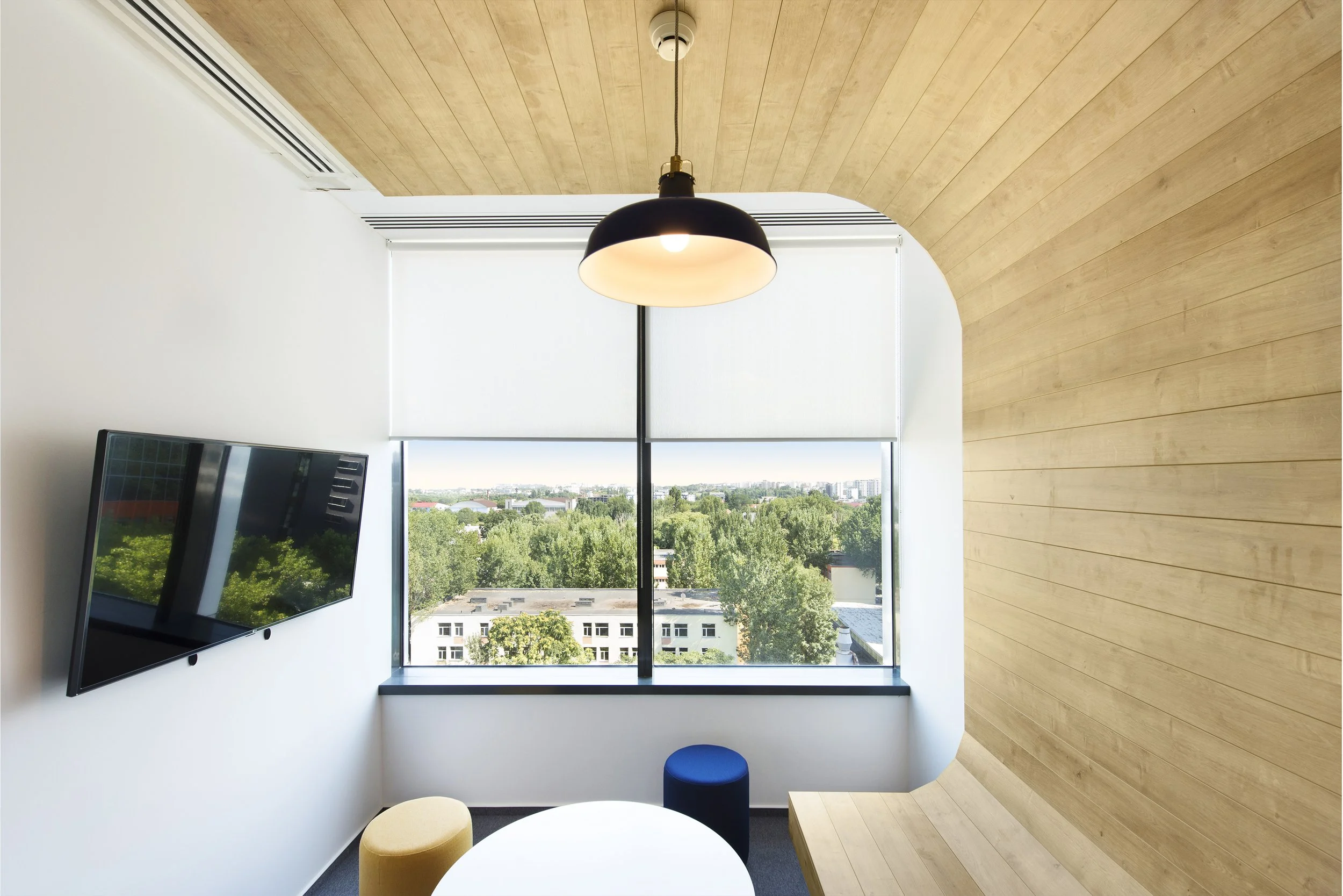 Modern office space with a large window showing a city view, a wall-mounted flat-screen TV, a hanging light fixture, and colorful round stools around a white table, with a wooden accent wall.