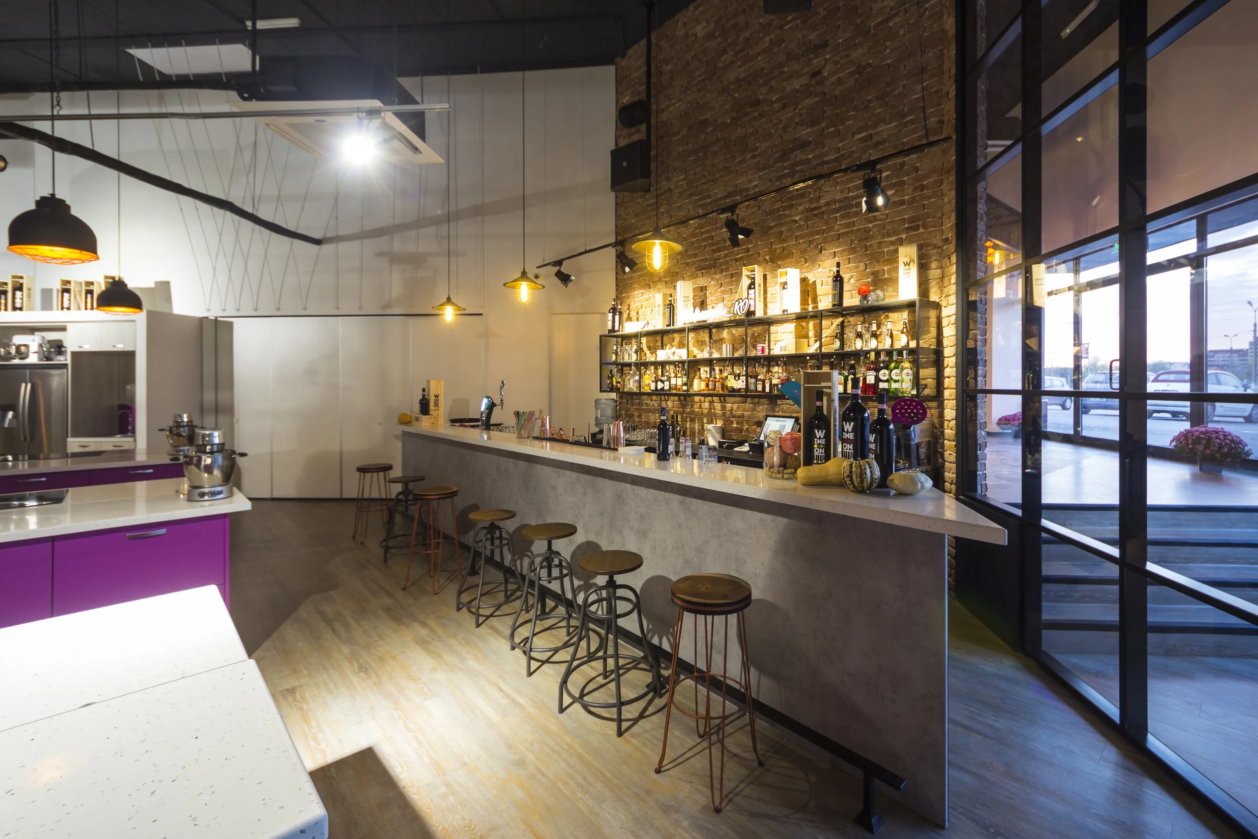 Interior of a modern cafe or bar with a long counter, barstools, a brick wall with shelves holding bottles, and large windows showing an outside parking lot.
