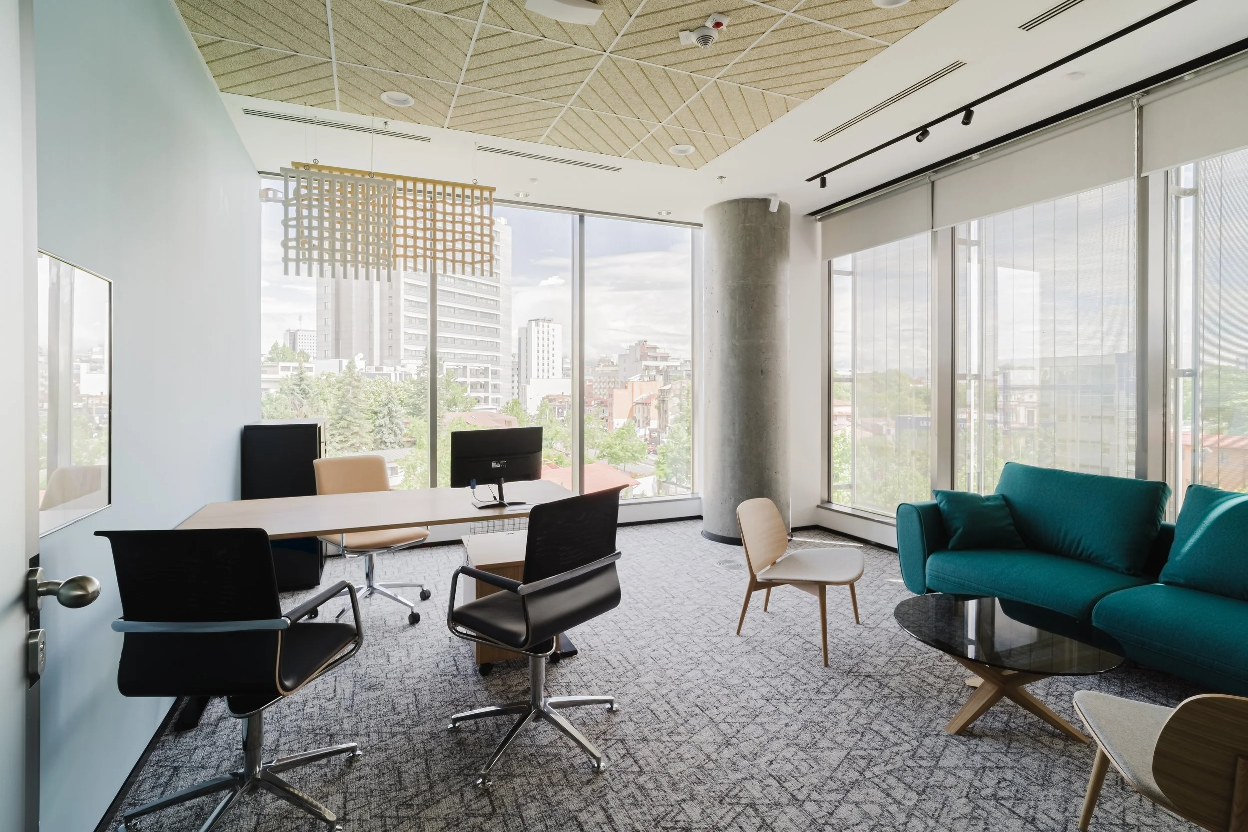 Modern office with large windows, a teal sofa, a wooden coffee table, two beige chairs, a black desk with a computer monitor, and black office chairs. City buildings and trees are visible outside.