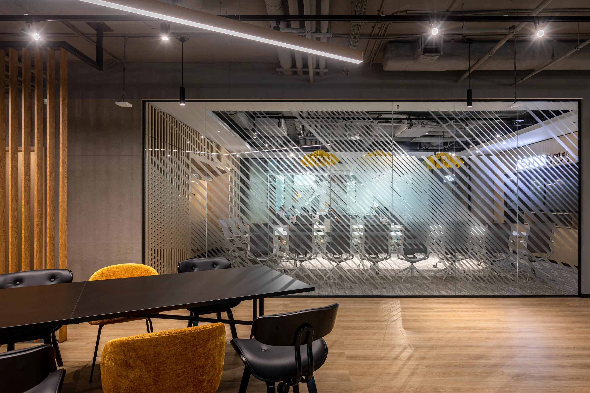Modern office conference room with glass wall, black chairs, and a wooden table, overlooking a workspace with additional chairs and tables.