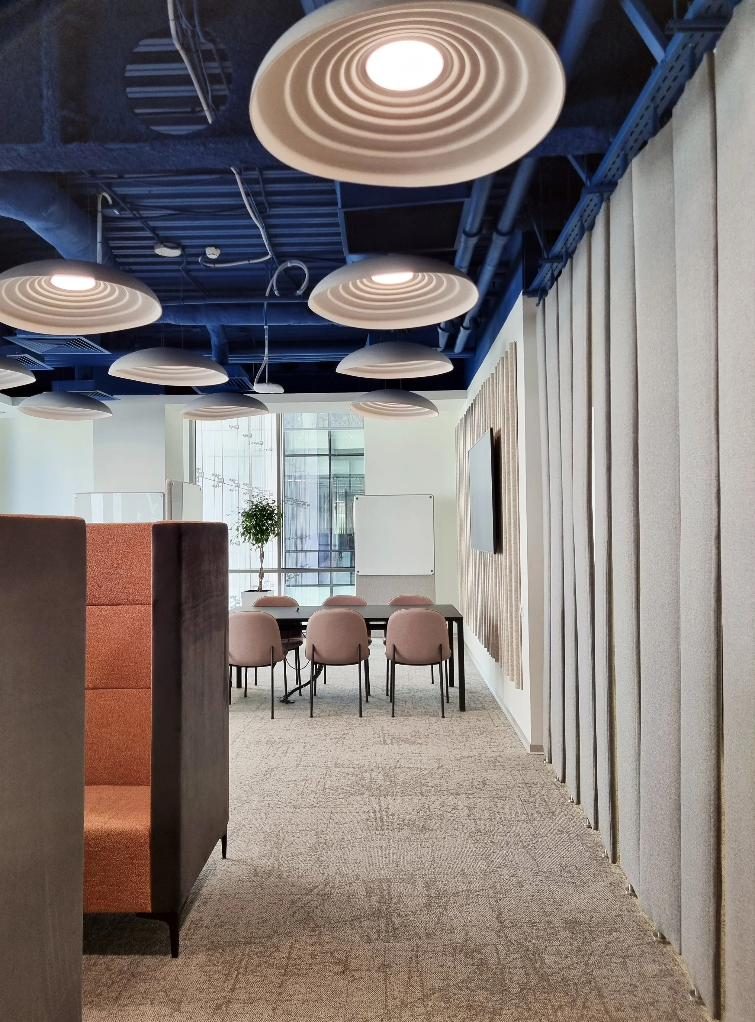 Modern conference room with beige chairs around a table, a whiteboard, a potted plant, large windows, and ceiling lights.