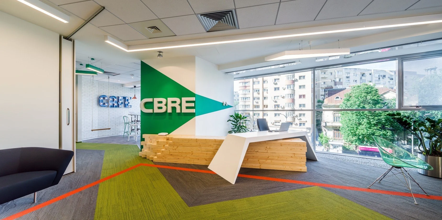 Modern office reception area with CBRE branding, a wooden desk, green and neutral colored carpet, large windows with city view, and minimalistic furniture.