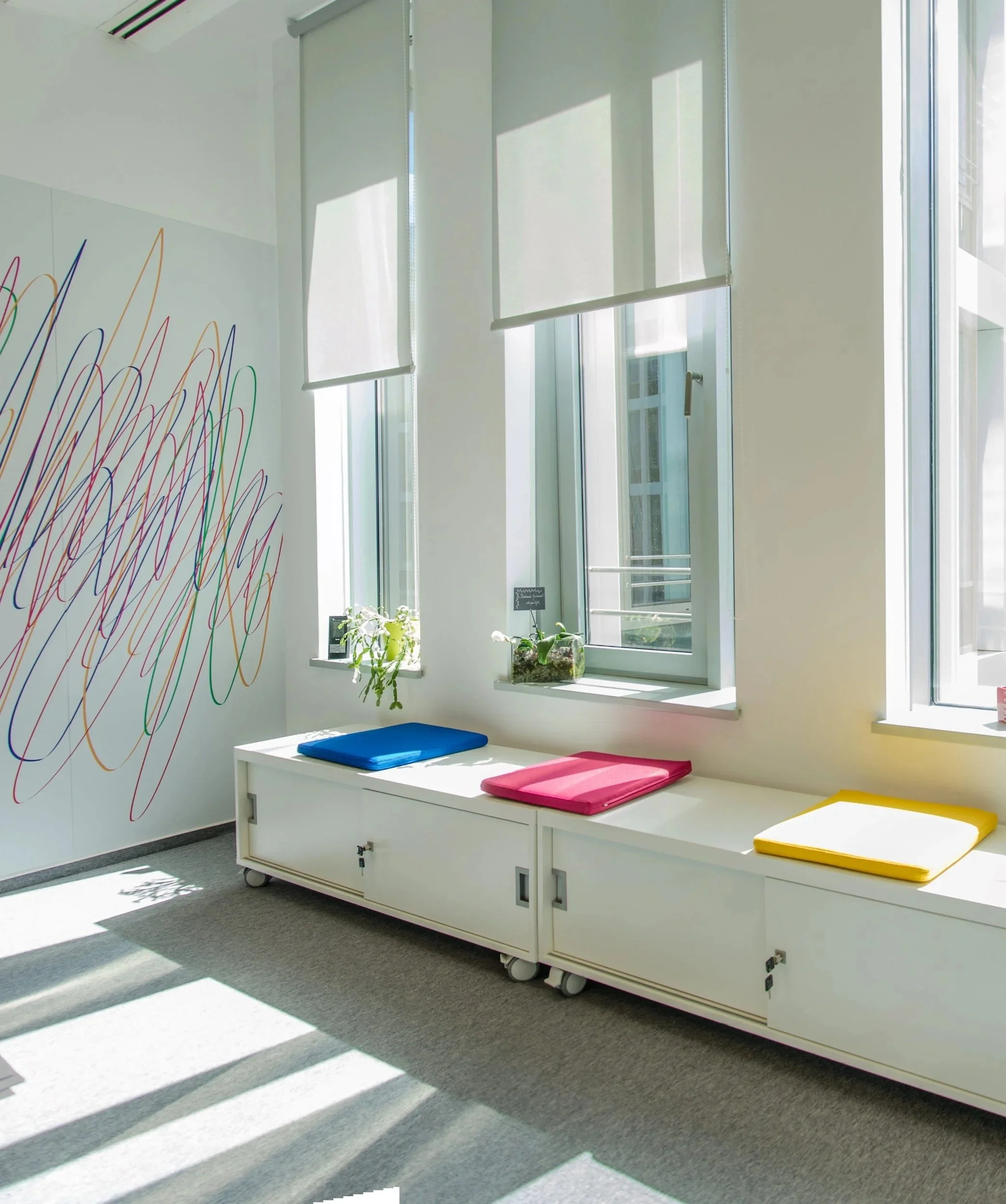A bright room with three large windows with white blinds, a white bench with colorful cushions, and a white wall with colorful abstract line art.