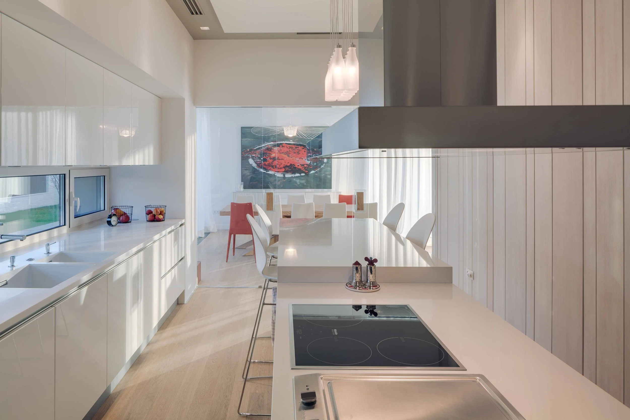 Modern kitchen with white countertops, light wood cabinets, a cooktop, and a dining area with white and coral chairs, artwork on the wall, and pendant lights.