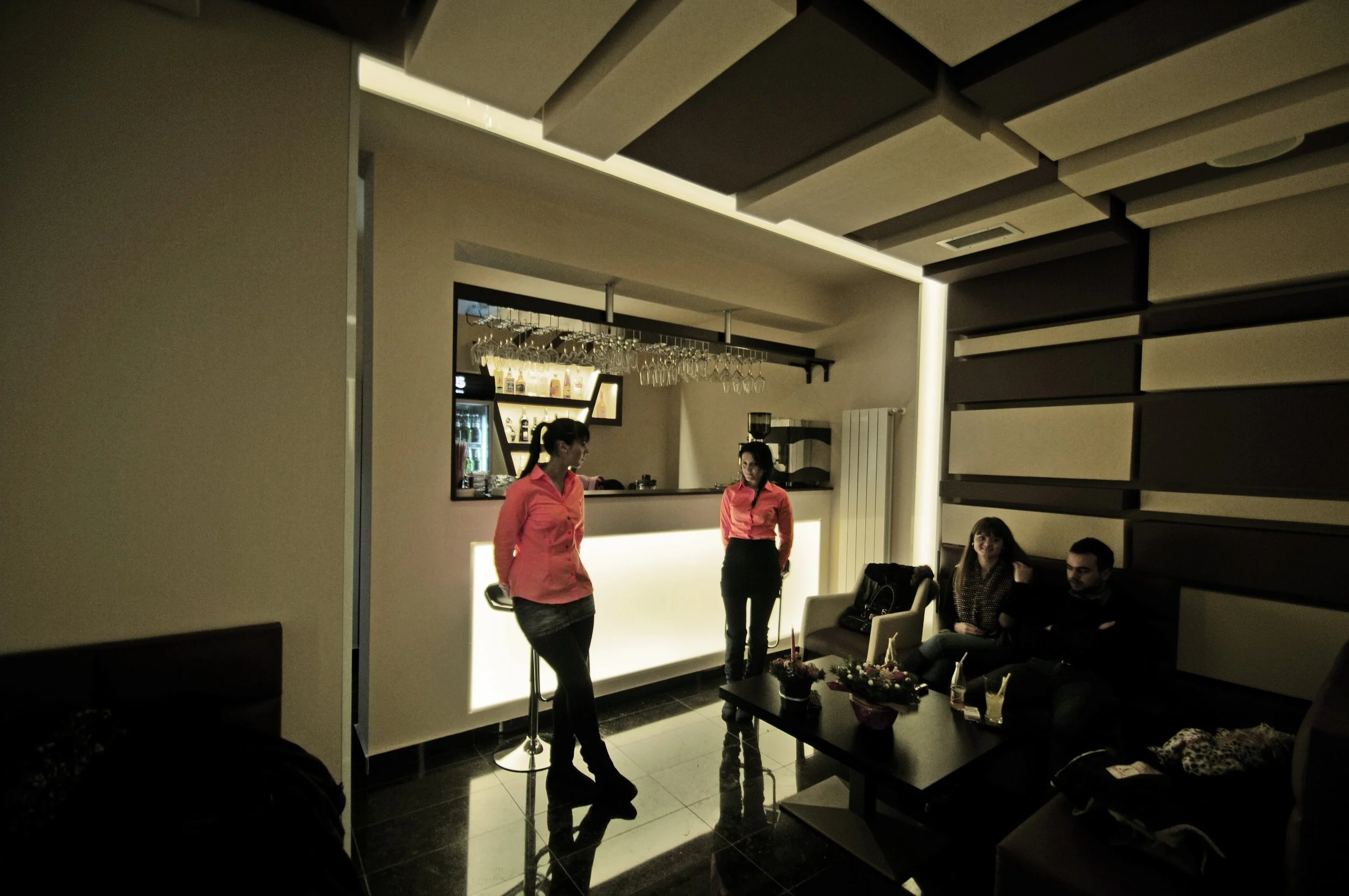 A dimly lit lounge area with two women in pink shirts standing near a bar, and a couple sitting on a sofa, with a table of flowers and drinks in front of them.
