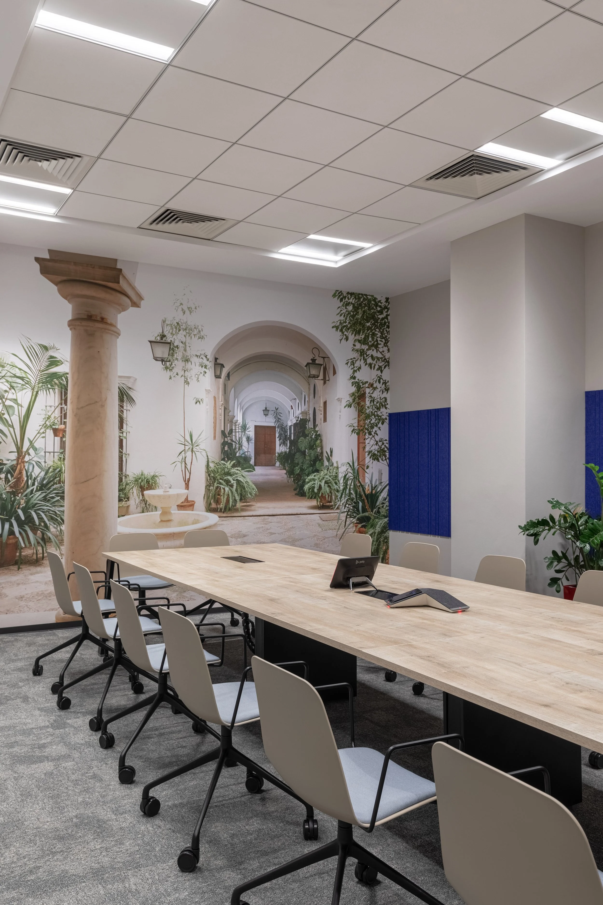 Modern conference room with a large wooden table, white chairs, and a mural of an outdoor corridor with plants and arches on the wall.