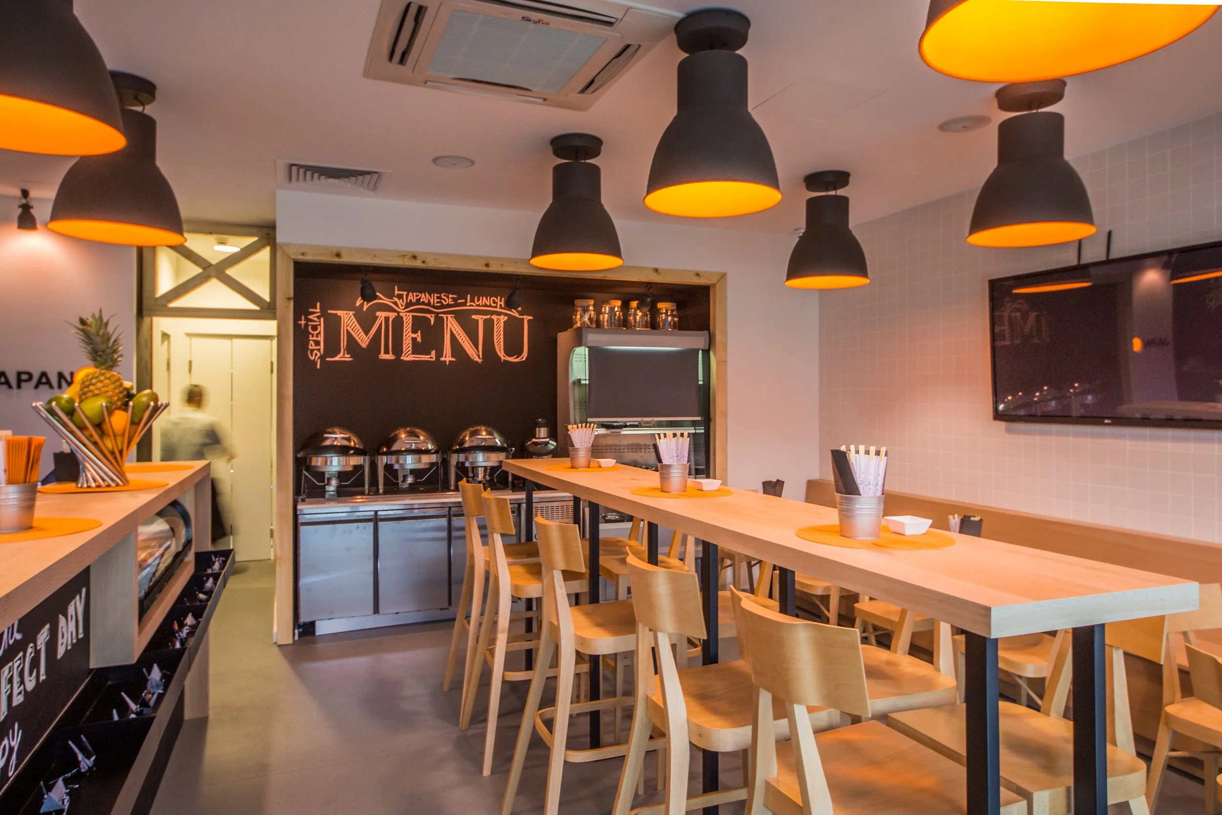 Interior of a modern Japanese restaurant featuring a long wooden high table with chairs, pendant lights with orange interiors, a blackboard menu with "Special Japanese Lunch Menu" written on it, and a fruit display with pineapples and other fruit on the left.