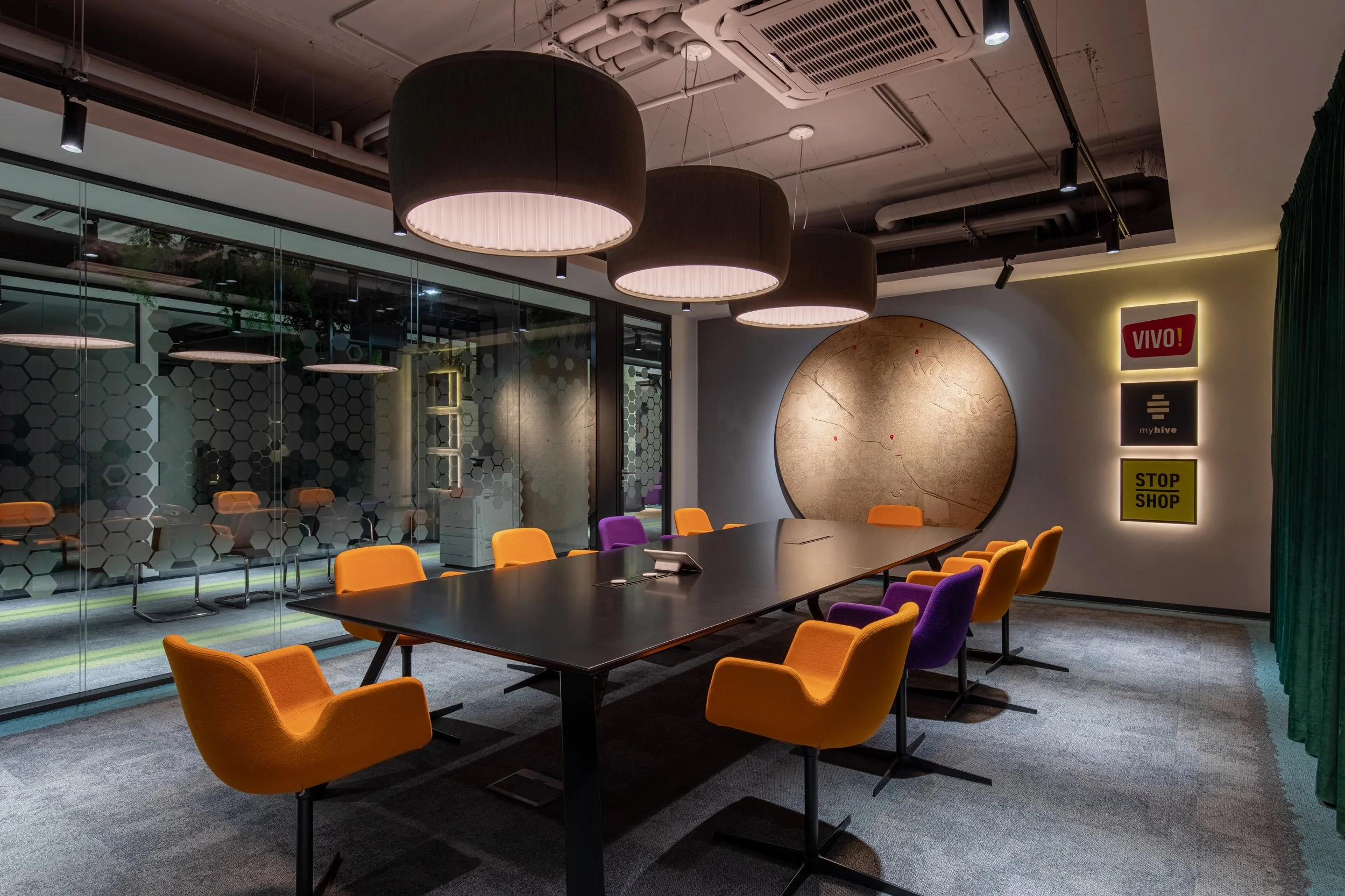 Modern conference room with a long black table, orange and purple chairs, large black ceiling lights, and a glass wall with honeycomb patterns. The wall features illuminated signs and a large textured circular artwork.