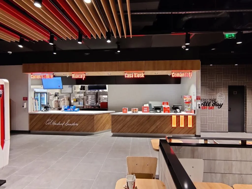 Fast food counter with a digital ordering screen, cash registers, and a kiosk, with signs in Romanian for ordering, paying, and the kiosk, inside a modern restaurant or fast food place.