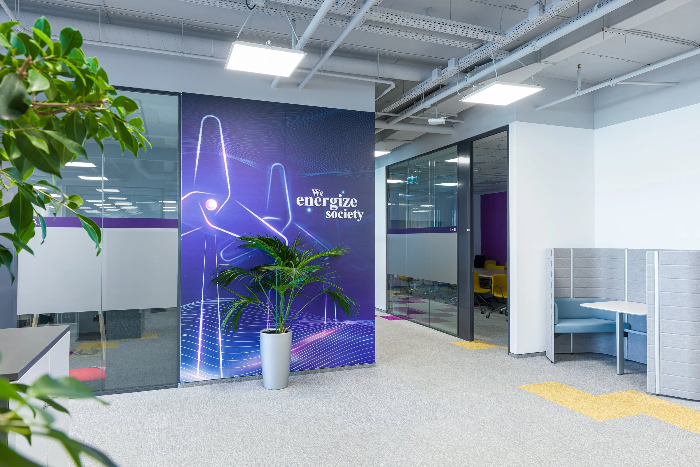 Modern office corridor with a large digital wall display reading 'We energize society,' potted plant, glass-walled meeting room with yellow chairs, and a photo booth with a gray booth and white table.