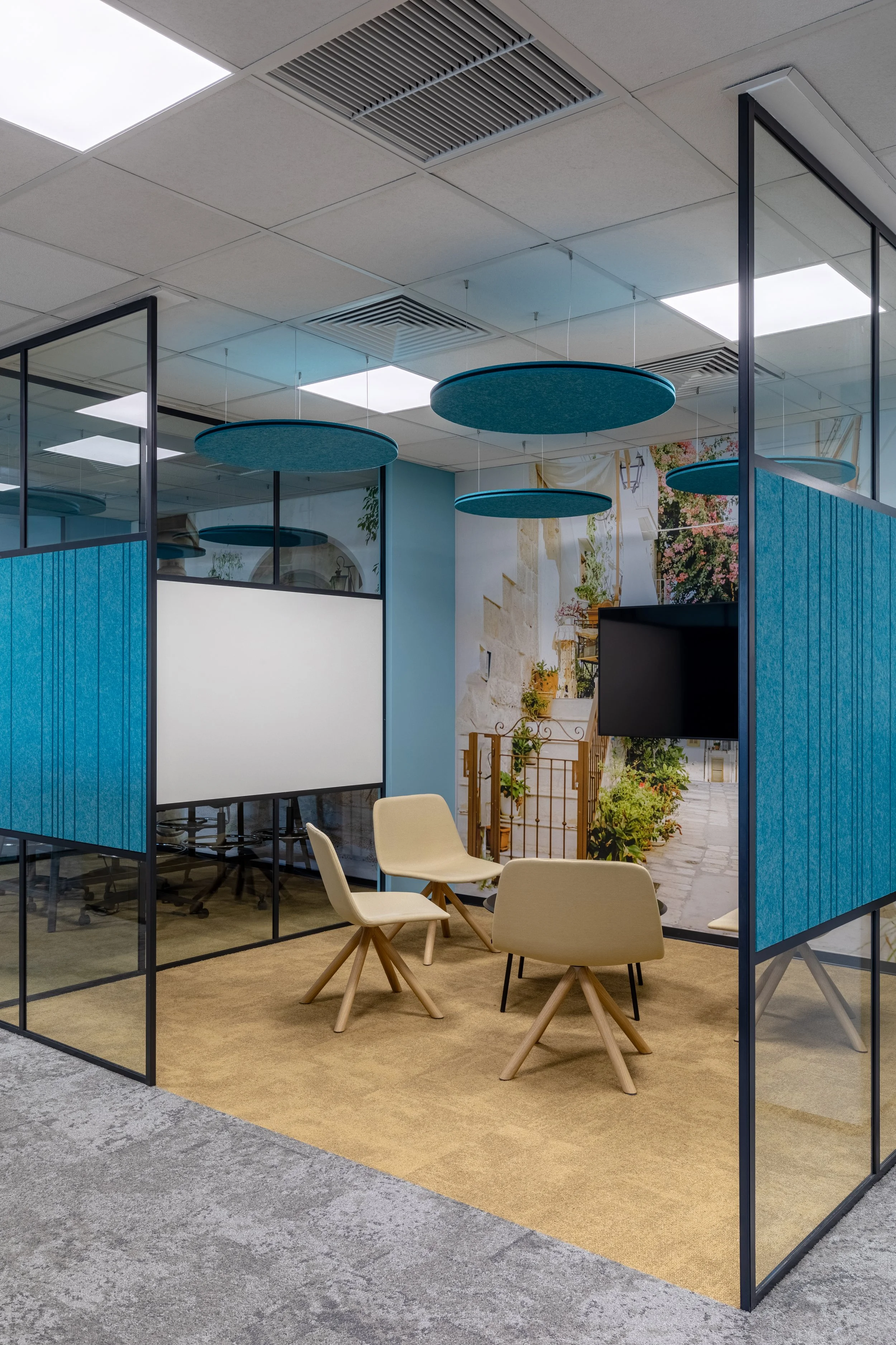 Modern office meeting room with four beige chairs, a large TV, glass walls, blue ceiling accents, and a mural of a Mediterranean street scene on the back wall.