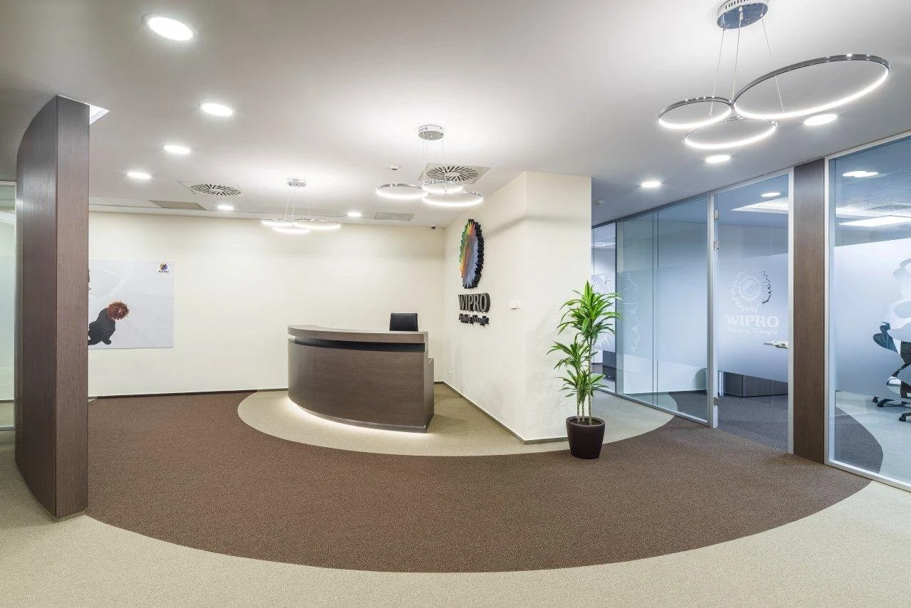 Modern office reception area with a wooden desk, a black chair, a potted plant, glass walls with frosted logo, and ceiling lights.
